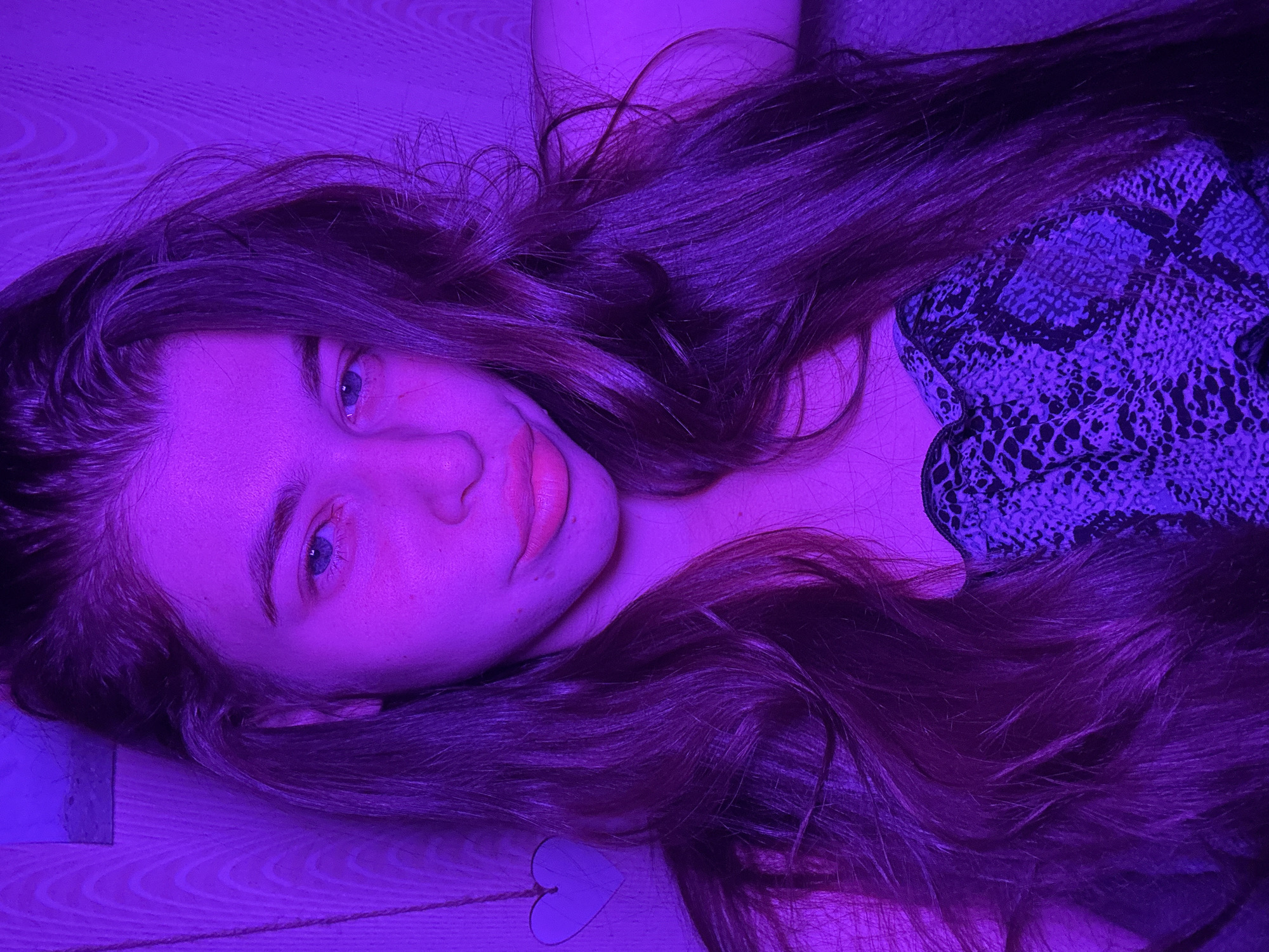 A person is lying on a wooden floor with long, dark hair spread around them. The person's face is visible, with light-colored eyes and lips. They are wearing a top with a snakeskin pattern. The scene is illuminated with a purple light. A small, white, heart-shaped object is on the floor near the person's hair.
The location is not apparent from the image. It is unlikely to be possible to determine if the scene is in the city of Мещёры, Russia, or anywhere else.