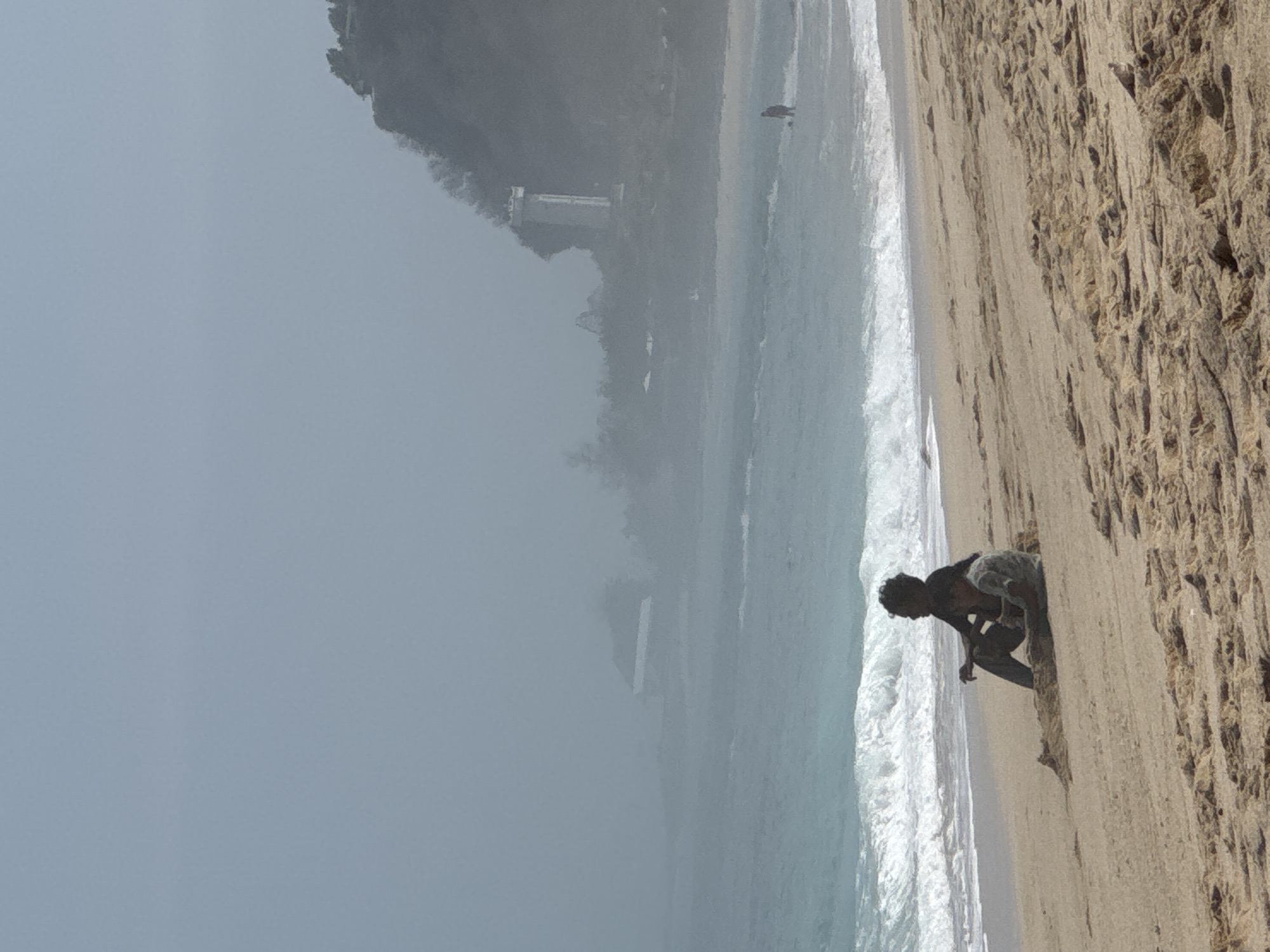 In Kuta Selatan, Indonesia, two people sit on a sandy beach with the ocean in the background. The person on the left, appearing to be an adult, is seated with their legs crossed and is looking towards the right. To their right and slightly in front, a child is seated, also facing towards the right. The waves of the ocean are breaking on the shore behind them. In the distance, a tall, white structure, possibly a lighthouse, is visible on a rocky, tree-covered hillside. The sky is overcast and appears hazy. A solitary figure is seen standing in the water further down the beach. The sand is textured with ripples and small debris.