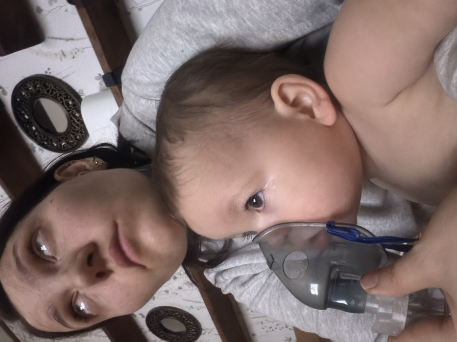 A woman lying down next to a baby. The baby is facing the camera, wearing a nebulizer mask. The woman's head is tilted to the side, and she is looking up at the camera. The background is blurry but appears to be a bed with a patterned sheet. Two decorative, round, dark objects are on the bed. The baby's skin appears light-colored, and one of their arms is visible. The woman has dark hair pulled back, and earrings are visible.