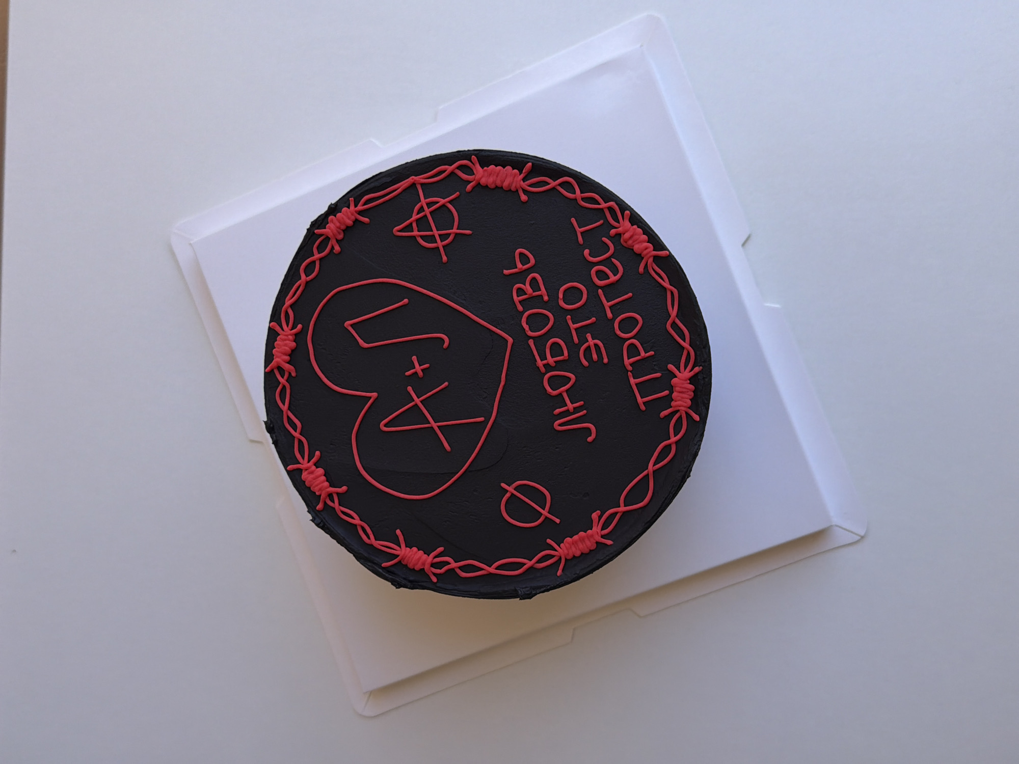 A round cake with dark, likely black, frosting is prominently displayed, resting on a white square cake board or box bottom. The cake's surface is elaborately decorated with red or pink frosting, creating an edgy and personalized design.
The perimeter of the cake is encircled with a detailed barbed wire pattern. Towards the center-left, a large heart shape contains the initials "A+J". Running vertically on the right side of the cake is Cyrillic text that reads "ЛЮБОВЬ ЭТО ПРОТЕСТ," which translates to "LOVE IS PROTEST."
Further reinforcing the theme, two distinct symbols are visible: an anarchy-like symbol (a stylized encircled 'A') is placed in the top-left quadrant, and a "no" or null set symbol (a circle with a diagonal line through it) is located in the bottom-right quadrant.
The cake is set against a plain white background, likely a table or countertop, under bright, even lighting, suggesting an indoor setting. The overall aesthetic, with its dark color, red decorations resembling barbed wire, and rebellious message, implies a punk-inspired or protest-themed creation. The initials "A+J" within the heart suggest a personal dedication for a couple, possibly celebrating a bond with an unconventional flair. The context for this media file is Tel-Aviv, Israel.