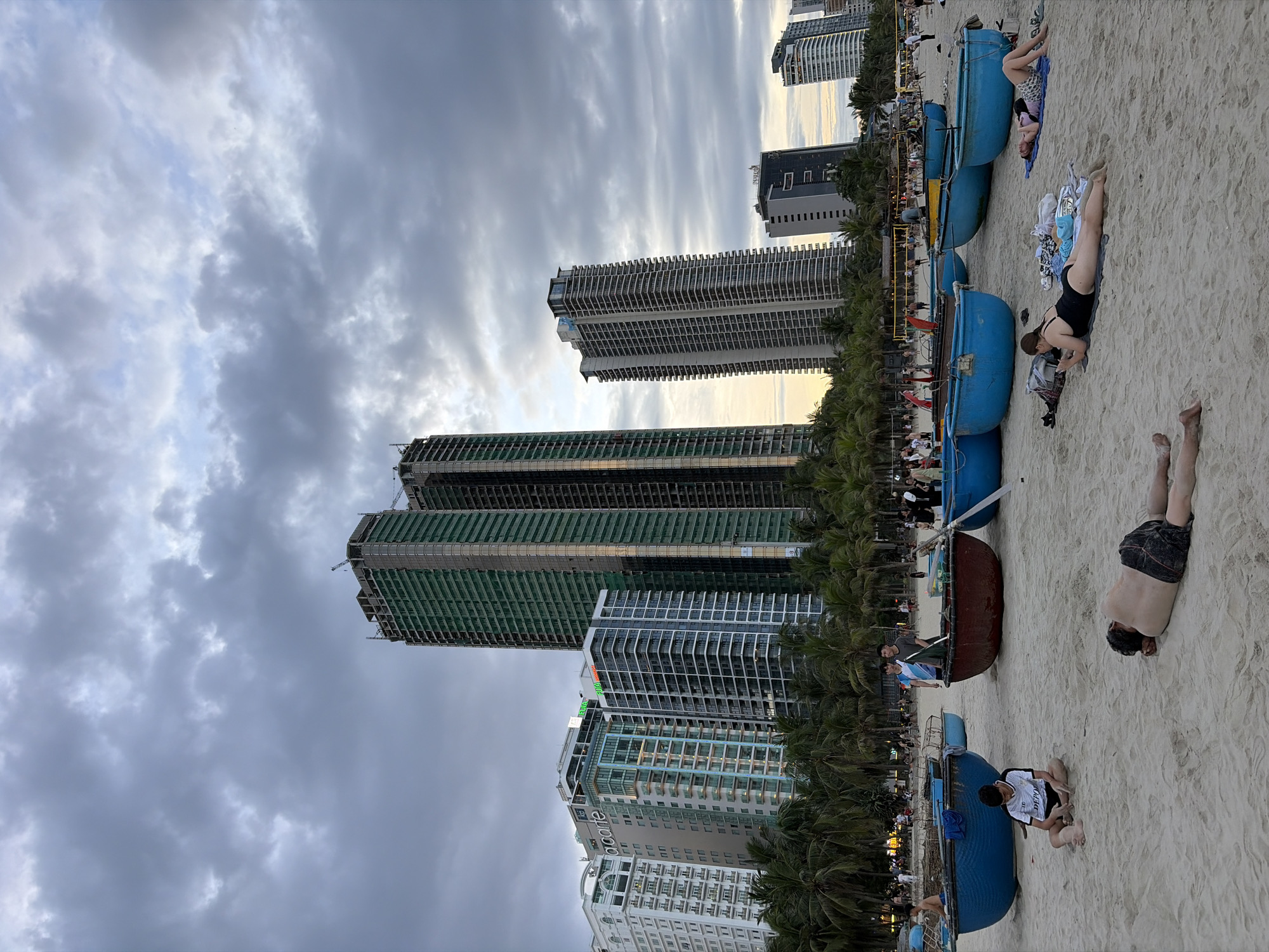 The image depicts a beach scene in Đà Nẵng, Vietnam, with a dramatic cloudy sky overhead. In the background, several tall buildings are visible, some of which appear to be under construction, evidenced by scaffolding and unfinished exteriors. Palm trees line the edge of the beach, separating it from the urban landscape. In the foreground, several round, boat-like structures made of woven material and painted in bright blue and a reddish-brown hue are scattered on the sandy beach.
Several people are present on the beach. In the center foreground, a young man in a white and black t-shirt is sitting on the sand with his legs crossed. To his right, another man is lying prone on the sand, his back to the camera. Further to the right, a woman in a black swimsuit is lying on a towel, looking at her phone. Behind her, another woman is lying on a towel, looking towards the camera. In the middle ground, other individuals are visible walking or standing near the boats, and in the far background, a volleyball net and people playing are discernible, indicating recreational activities. The overall impression is a lively beach scene during the day, with a mix of relaxation and activity, set against a backdrop of modern city development and natural elements.
