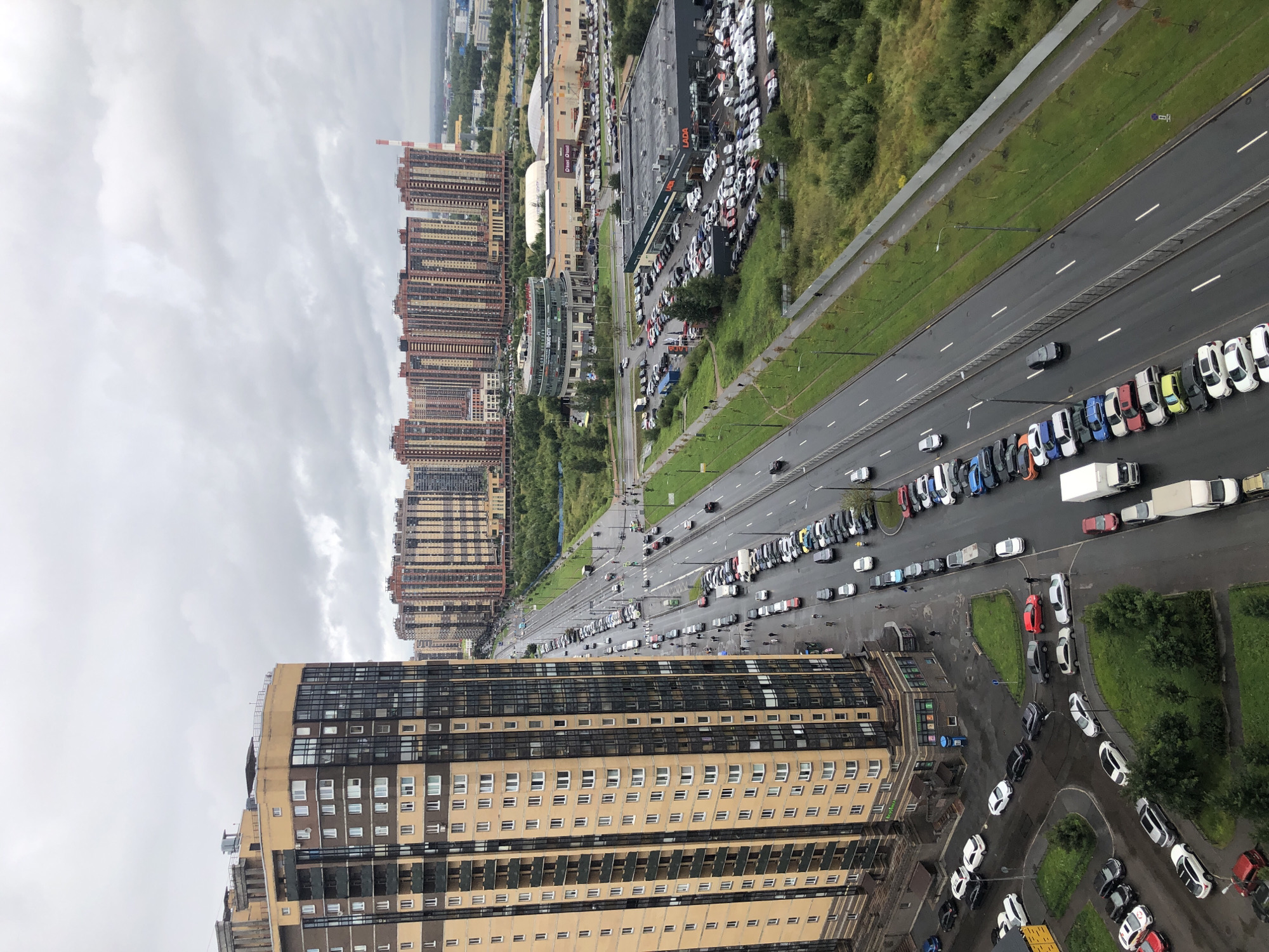 An aerial, eye-level medium shot captures a wide, multi-lane road with moderate traffic and parked cars on the right, flanked by lush green trees and a white metal fence on the right side of the road. On the left of the road, a tall, tan and dark brown apartment building dominates the foreground. Beyond the road, rows of uniform, tan and brown high-rise apartment buildings stretch into the distance under an overcast, grey sky. The road itself is wet, indicating recent rain. Cars are visible on both sides of the road, with some parked and others in motion. Several pedestrians can be seen walking along the sidewalks. To the right of the apartment building, a car dealership with a LADA sign and many cars parked outside is visible. Further back, a circular building with a glass facade and several rectangular shopping centers with parking lots are situated. The overall scene depicts a modern urban environment with high-rise residential buildings, commercial areas, and extensive road infrastructure.