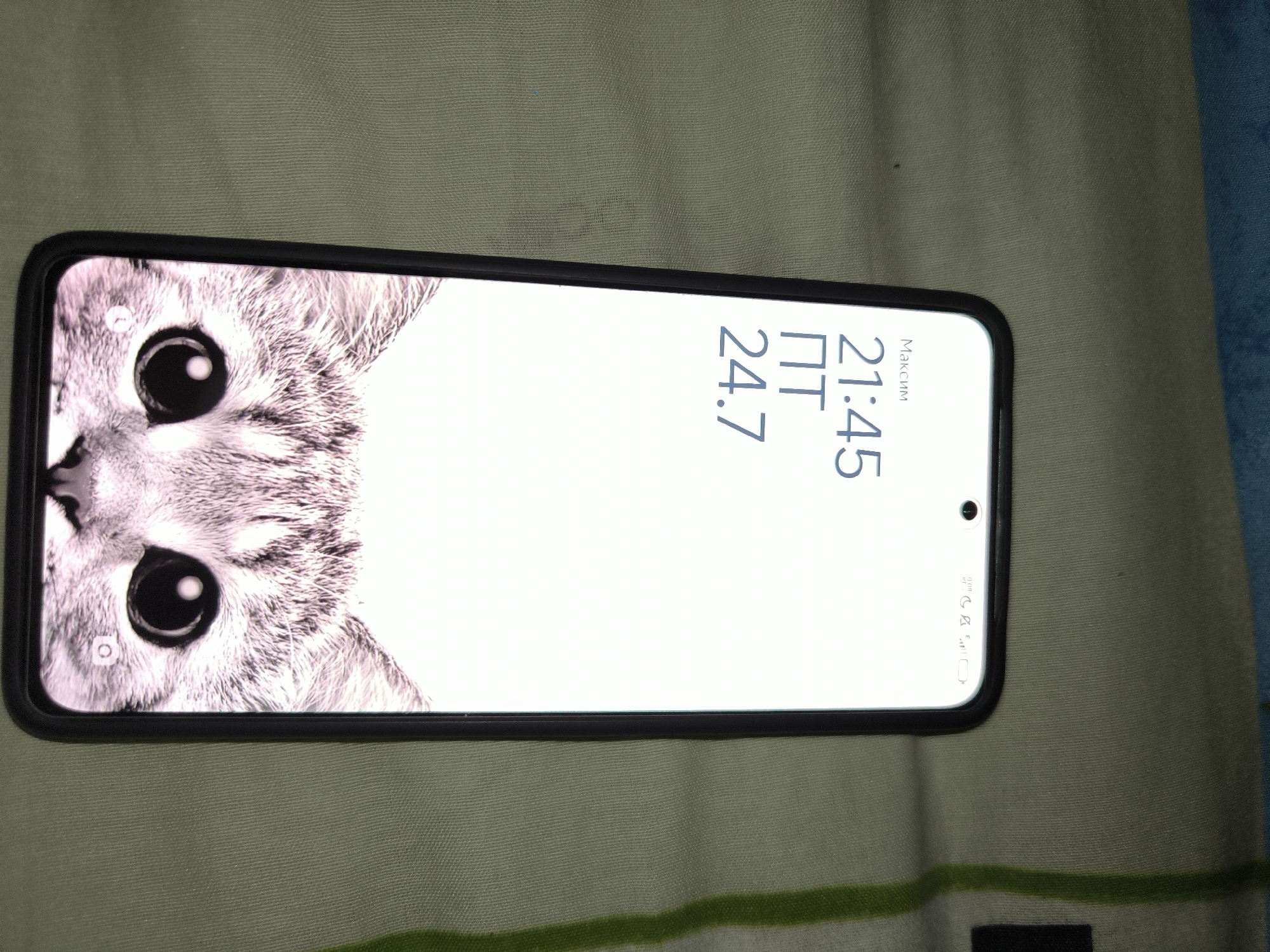 The image shows a smartphone lying on a textured, olive-green surface. The phone displays a white screen with a wallpaper of a cat's face on the left side. The time is shown as "21:45" and the date as "24.7" on the right side, with the word "Максим" above the time. The phone is oriented horizontally, with the top edge towards the left. The surface beneath the phone appears to be fabric with a subtle weave pattern, and there are faint green stripes running horizontally across it. To the right of the phone, a sliver of a blue textured surface is visible. No people or animals are visible in the image other than the cat on the phone's wallpaper. The location is not discernible from the image.