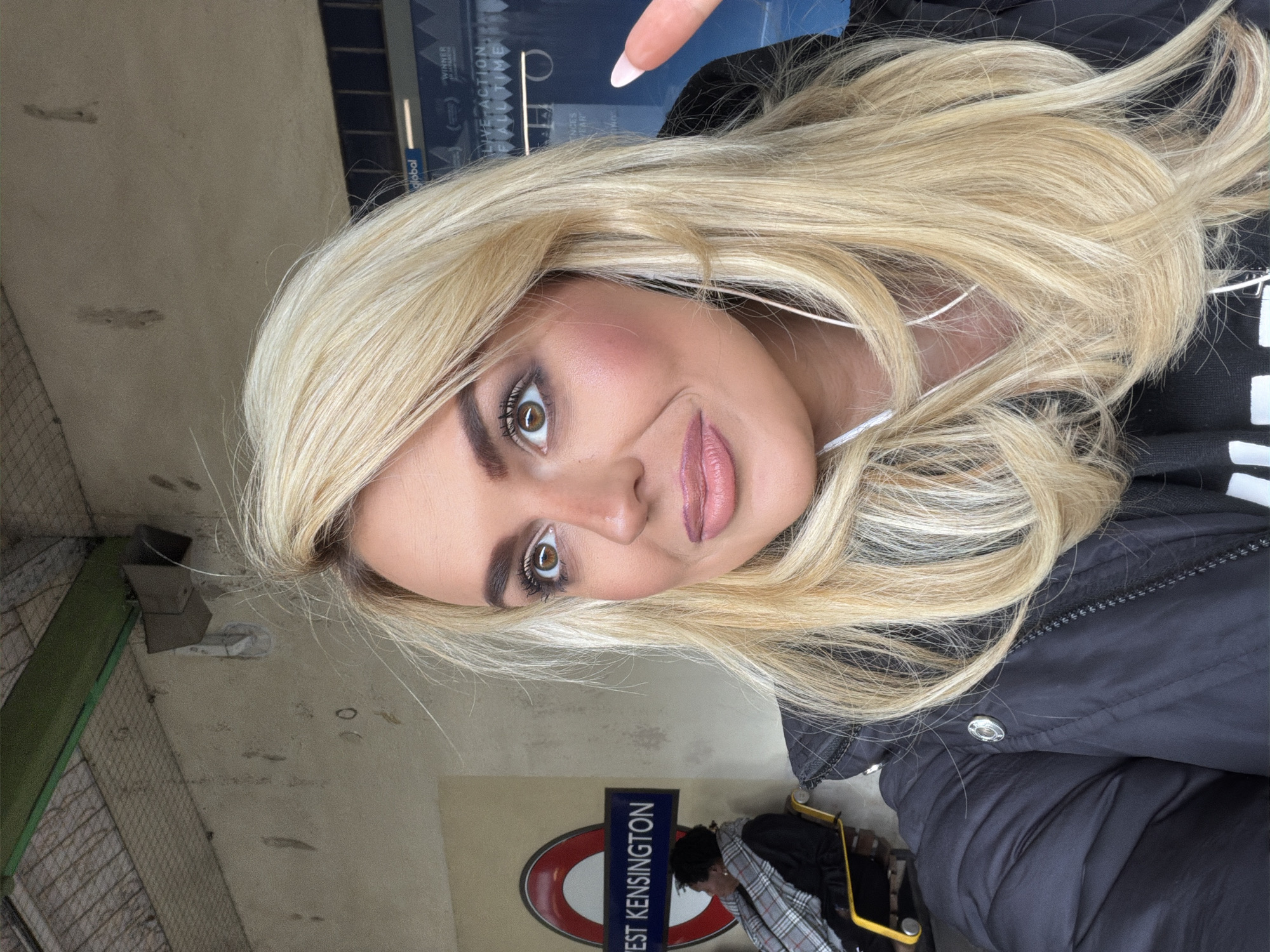 A woman with long blonde hair is taking a selfie in front of a West Kensington London Underground station sign. The sign is a red circle with a blue rectangle in the center with the text "WEST KENSINGTON" in white. Another person is visible slightly obscured behind the woman. The wall behind the sign is beige. The woman is wearing a black jacket. She is holding up a finger to the right of the frame.