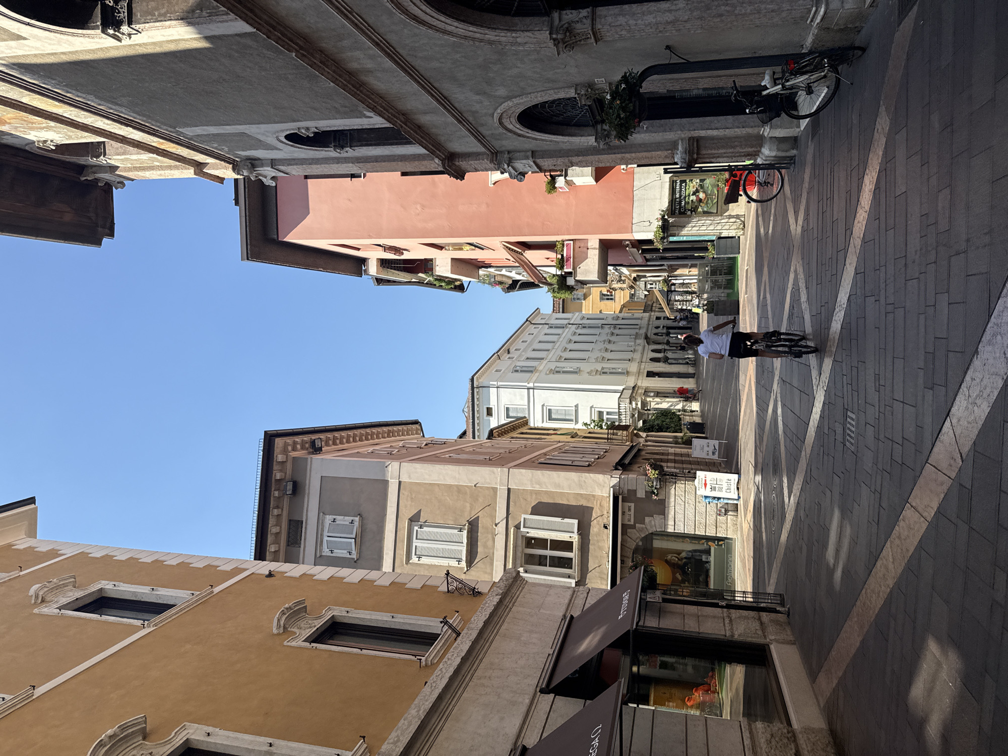 The image depicts a view down a narrow street in Trento, Italy, likely in the historic center given the architecture. The buildings lining the street are multi-story structures with various architectural styles, exhibiting different colored facades and window designs. On the left, a building with a mustard-yellow facade has decorative window frames and a dark awning over a shop entrance. To its right, a lighter-colored building features multiple windows with white shutters. Further down the street, a vibrant pink building stands out. On the right side of the street, a building with a rough, greyish facade features arched openings, a decorative cornice, and window planters.
The street itself is paved with dark cobblestones, marked with lighter diagonal and intersecting lines. Sunlight casts shadows from the buildings, creating a textured pattern on the ground. A person on a bicycle is in the middle of the street, moving away from the camera, wearing a white t-shirt and dark shorts. Several bicycles are parked along the right side of the street. Signs are visible on some of the shop fronts and on the street, indicating commercial activity. The sky is clear and blue, suggesting a sunny day. The overall impression is of a charming, sun-drenched European street scene.
