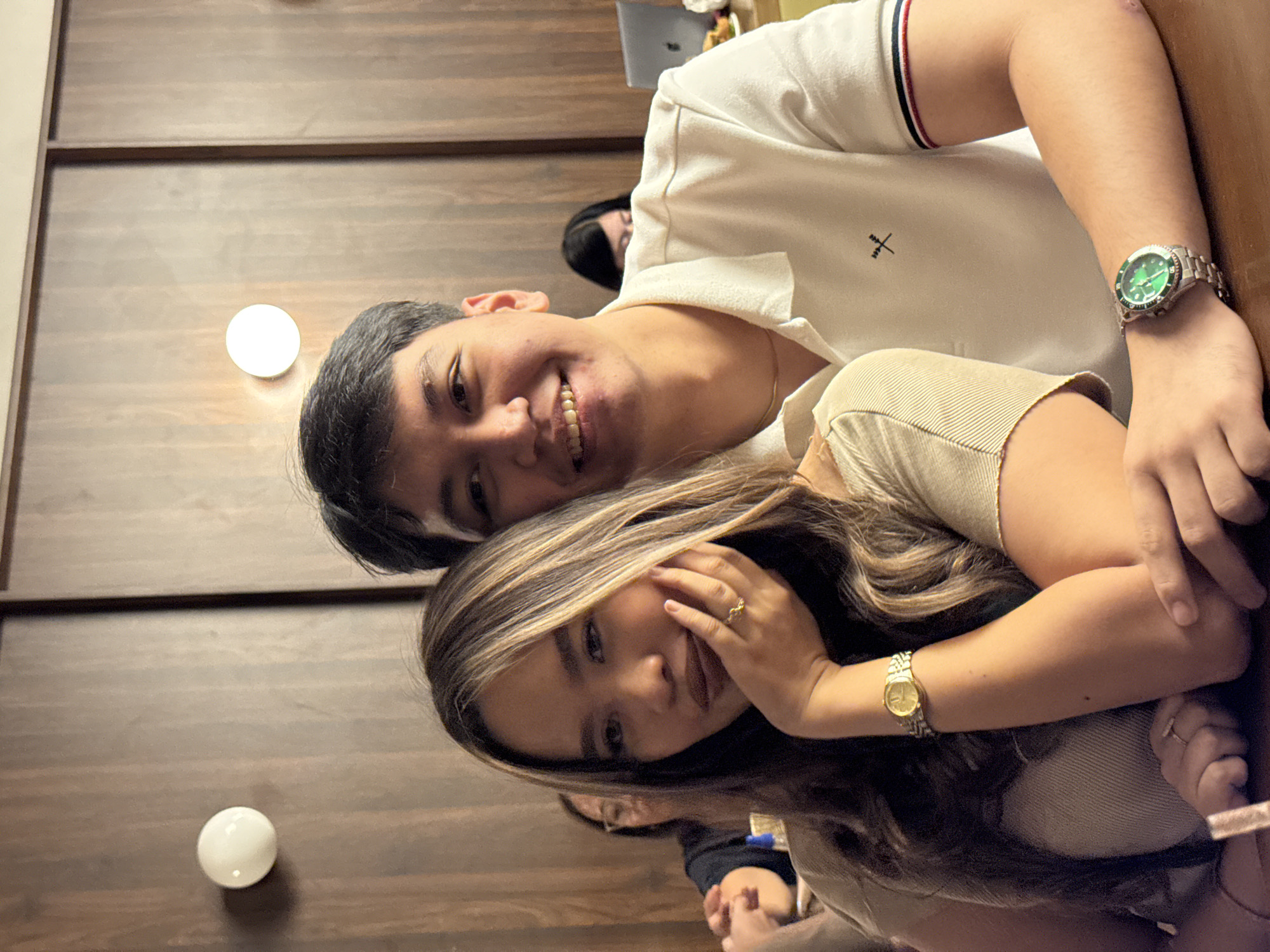 Two people are posing closely for a photo. The man has short black hair, a white polo shirt with red and navy stripes on the sleeve cuff, and a silver watch with a green face. He is smiling, and part of his gold necklace is visible. The woman has long brown hair with blonde highlights and is wearing a light brown top. She is smiling and has a gold watch on her wrist and a ring on her finger. The background shows a wooden wall with two spherical lights. In the background, other people are partially visible. The setting could be a restaurant or indoor gathering in Davao City, Philippines.