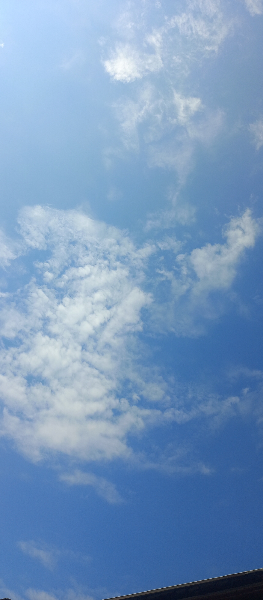 A clear blue sky with scattered white clouds fills most of the frame. The clouds are wispy and varied in density, with some appearing brighter and more defined, suggesting sunlight filtering through. A dark, horizontal object, possibly a roof or building ledge, occupies the very bottom of the image, suggesting this is an outdoor scene viewed from a lower elevation looking upwards. The lighting indicates it is daytime, likely sunny. There are no people or animals visible in the image, and no discernable urban elements besides the partial structure at the bottom. The location in Голуха, Russia, is noted, but the image itself provides no specific geographical or architectural clues to confirm this.