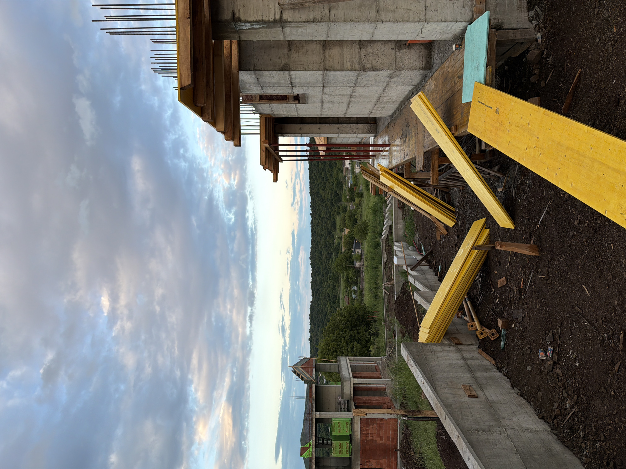 The image shows a construction site located in Cherni Vrah, Bulgaria, with an unfinished concrete structure in the foreground. Exposed rebar extends from the top of the concrete. Yellow wooden planks are scattered nearby on the ground, which appears to be damp soil. In the background, there's a view of a rural landscape with houses and trees. The sky is cloudy with hints of light.