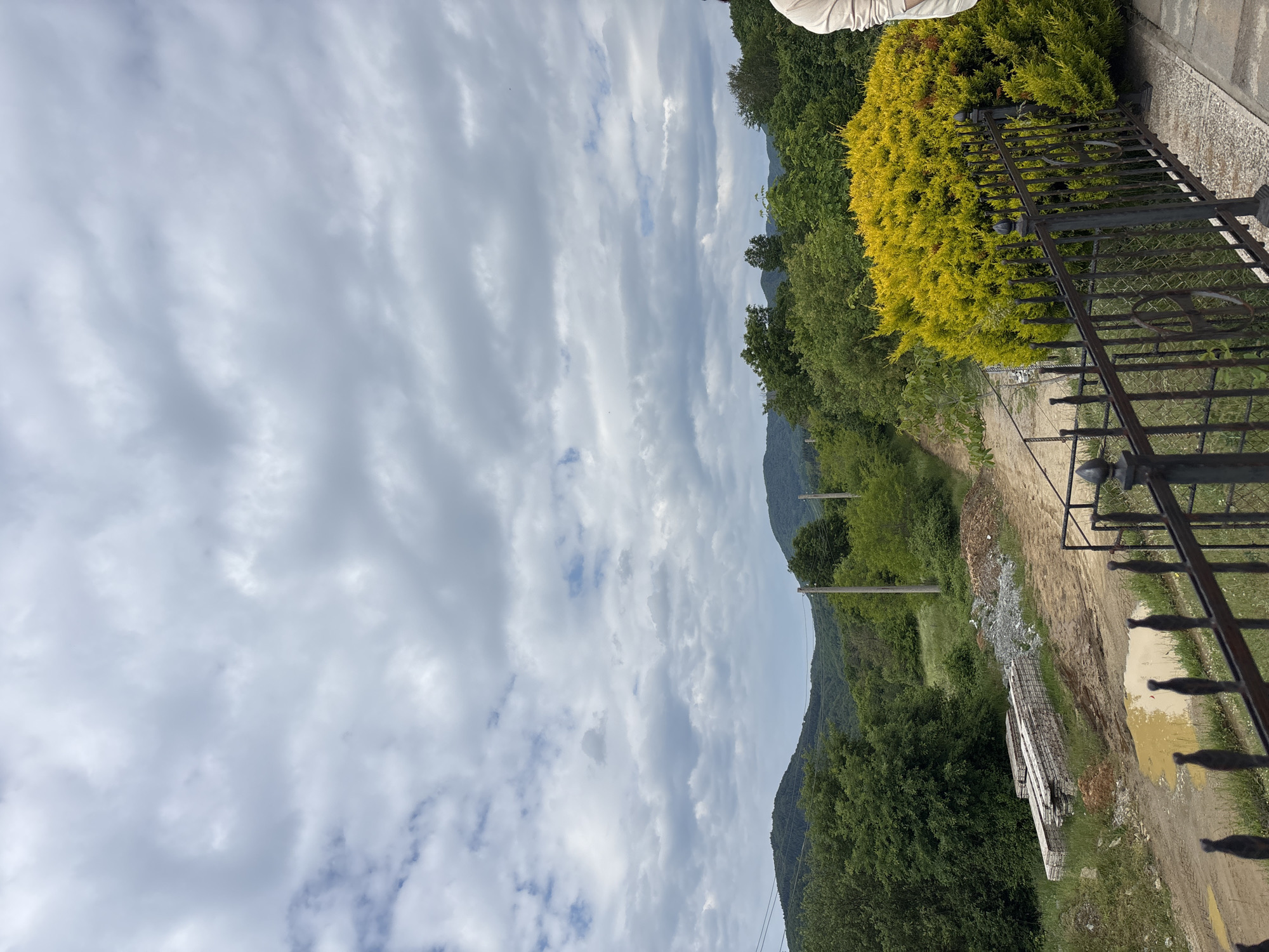 This outdoor image depicts a rural scene, possibly in the vicinity of Zlatograd, Bulgaria, though no specific city landmarks are visible. The foreground features a portion of a black, wrought-iron fence and some low-lying evergreen shrubs with golden foliage. Beyond the fence, a rough, unpaved path or road stretches into the distance, flanked by dense green vegetation and trees. The terrain ascends slightly toward a ridge or small mountain visible in the midground. Overhead, a cloudy sky dominates the upper portion of the frame. Construction materials, including what appear to be pre-fabricated concrete elements, are piled near the path, suggesting ongoing building or infrastructure work in the area. Electrical poles with power lines are also visible along the road. There are no people visible in the frame.