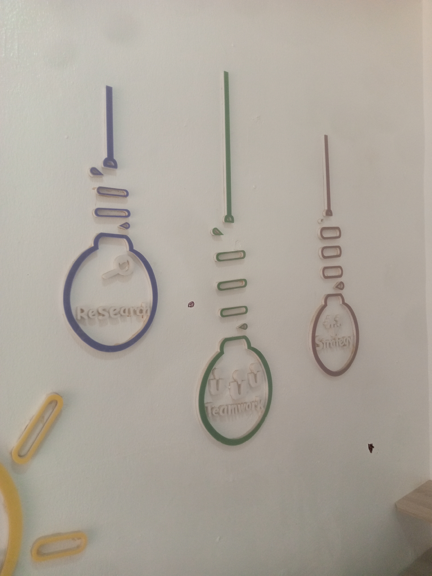 The image shows a white wall with three stylized light bulb shapes affixed to it. Each light bulb has a different color outline and a word inside the bulb: "Research" (blue), "Teamwork" (green), and "Strategy" (brown). Each light bulb is represented as an incomplete construction, with the top portion detached and segmented. Additionally, there are yellow shapes resembling segments of a larger object on the lower left side of the wall. The location is Abuja, Nigeria.