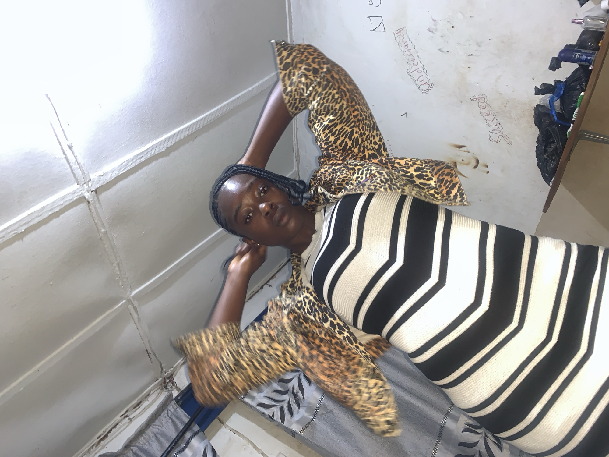 A young Black woman with braided hair stands in a dimly lit room, wearing a leopard-print jacket over a black and white striped dress. Her arms are raised behind her head, and she looks towards the camera with a neutral expression. The room appears to be a bedroom, with a patterned bedspread visible and a cluttered shelf in the background holding black trash bags, toothbrushes, and bottles. The walls are white and appear to have some scribbled writing and drawings on them, including the words "Success" and "Understand." The ceiling is made of corrugated metal panels. The scene appears to be domestic. The location is New Stadium, Nigeria.