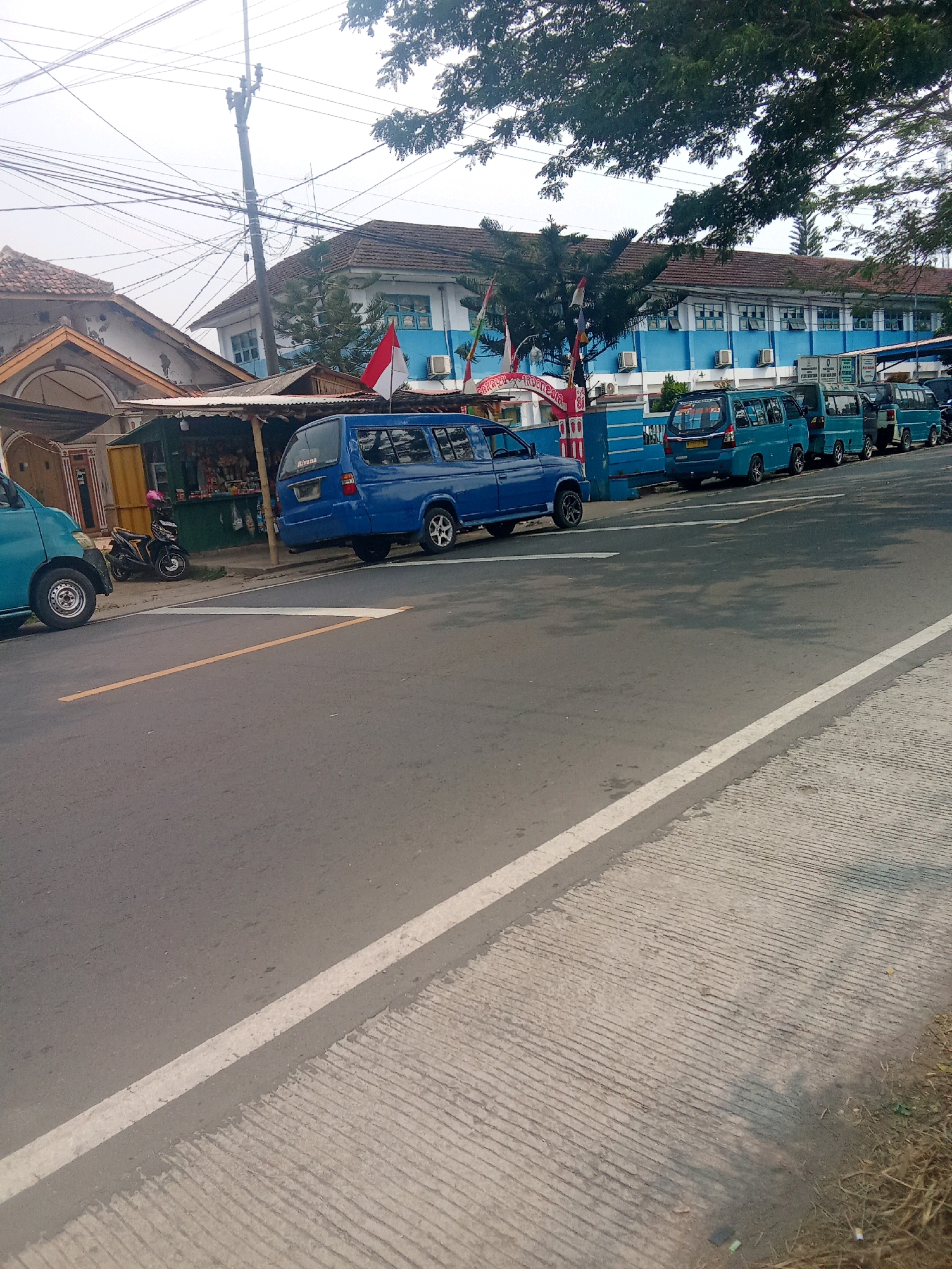 The image depicts a street scene in Warunggunung, Indonesia, with a blue building featuring multiple windows and air conditioning units dominating the background. Several blue vans are parked in a line along the side of the road in front of the building. To the left of the building, a smaller structure with a corrugated metal roof is visible, with a motorcycle parked in front of it. A blue pickup truck is parked on the left side of the road. The road itself is paved and has yellow and white lane markings. The foreground shows a portion of the road and a textured concrete sidewalk. Overhead, a complex network of electrical wires is strung across the sky, supported by a utility pole. Trees with green foliage are present, casting shadows on the road. The sky is overcast.