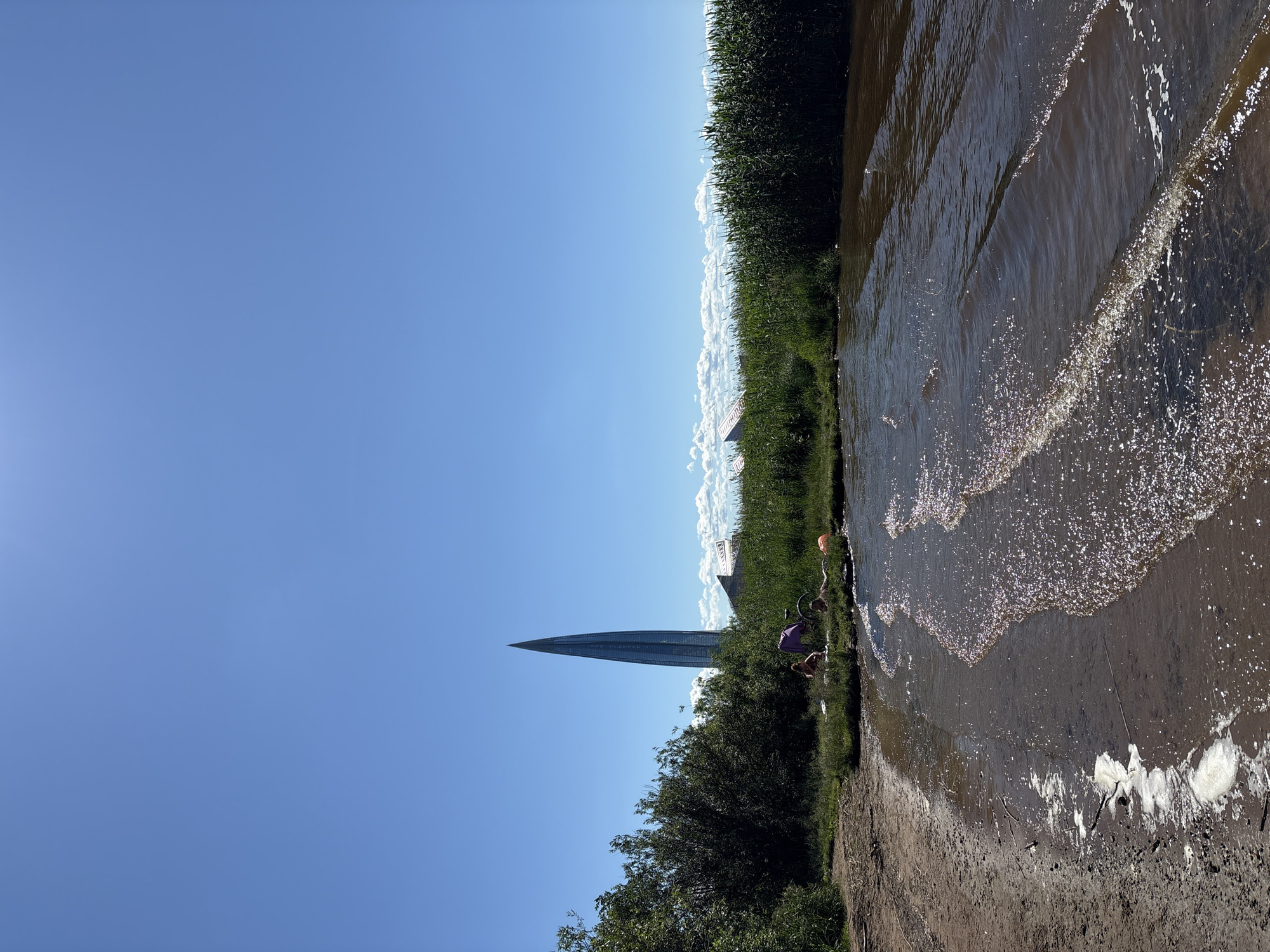The image depicts a sunny day at a body of water, likely a bay or river, in Saint Petersburg, Russia. In the background, the prominent Lakhta Center skyscraper, a distinctive modern architectural landmark, pierces the clear blue sky. Dense green reeds and bushes line the shore, with a few fluffy white clouds scattered near the horizon. On the grassy bank closer to the water, two individuals are visible, appearing to be relaxing. One person is sitting up, possibly engaged in an activity or conversation, while another person is lying down. A bicycle is parked nearby, along with a purple blanket and an orange object, suggesting a recreational outing. The water's surface is slightly rippled, with sunlight reflecting off the small waves and the sandy, somewhat muddy shore. The overall scene conveys a peaceful, outdoor setting with a backdrop of urban development.