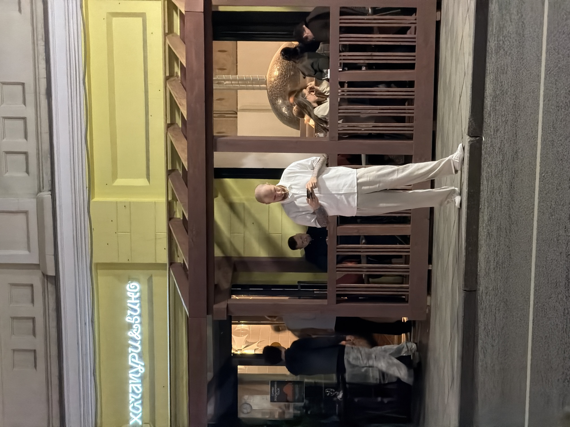 Here is a detailed description of the image:
**Scene**: The image appears to be an outdoor scene in what is likely a European city, due to the architecture and signage. Based on the presence of a neon sign in Cyrillic script reading "Хачапури&Вино" (Khachapuri & Wine) and details in the architecture, it is plausible that this is a view of a restaurant in Moscow, Russia.
**People**:
* A bald man wearing light-colored clothing is standing on the sidewalk next to a wooden structure. He is looking at a smartphone in his hands. Tattoos are visible on his forearms.
* Several people are visible inside the restaurant or bar, some seated. Their activities include conversing and dining.
* People are entering or leaving the establishment.
**Objects**:
* A wooden structure, possibly part of an outdoor seating area for the restaurant, is prominently visible.
* A large, shiny, globe-shaped object is visible inside the restaurant.
* A neon sign illuminates the storefront.
* A gray sidewalk abuts the building.
**Actions/Interactions**:
* The man is using his smartphone.
* People inside the restaurant are engaged in typical restaurant activities such as eating and talking.
**Environment**:
* The lighting suggests that it is evening or night.
* The architecture of the buildings is a mix of classical and contemporary styles.
* The setting is urban, with a restaurant or bar on a street.