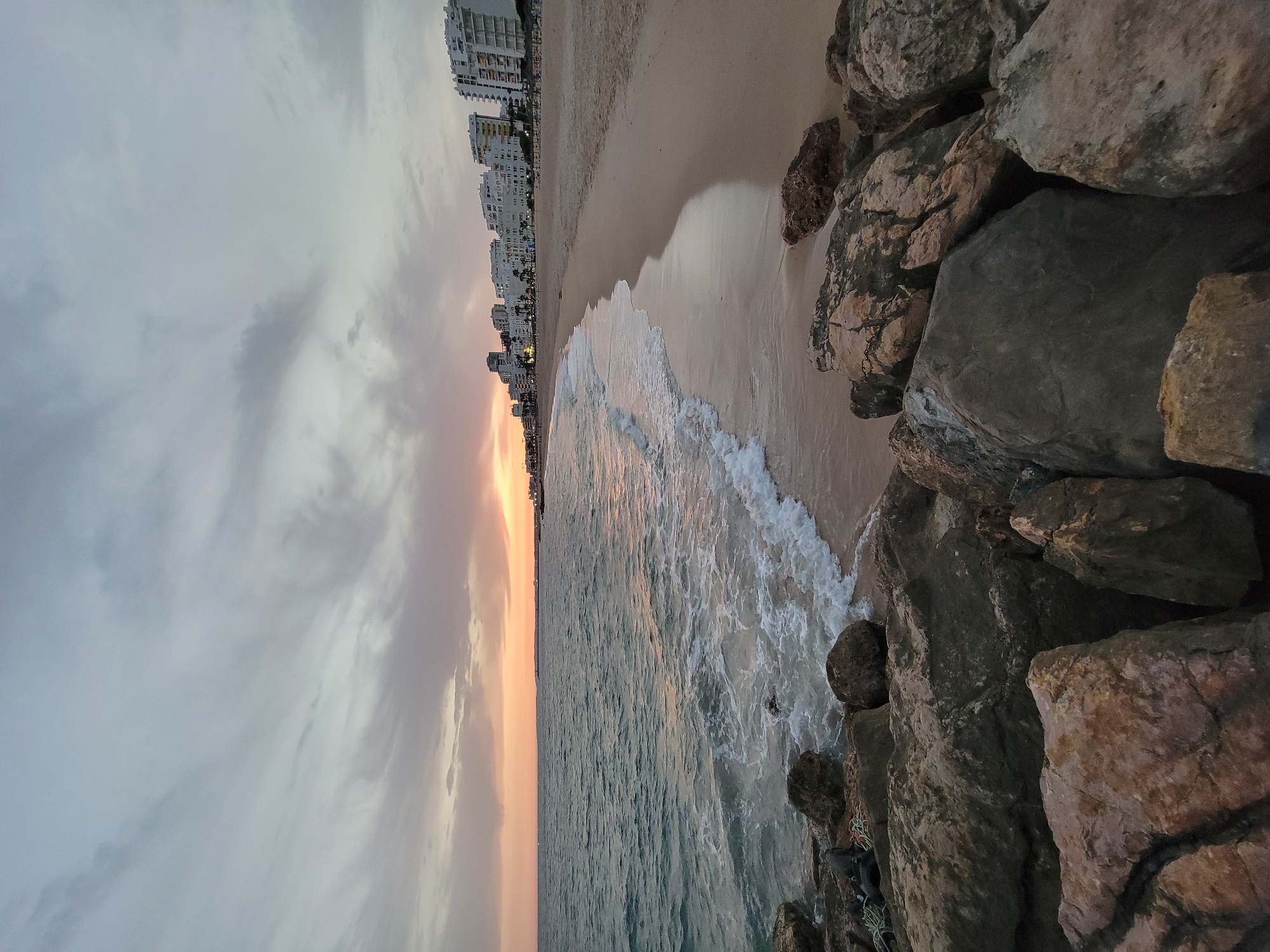 The image depicts a beach scene at dusk in Quarteira, Portugal. In the foreground, large, rough rocks line the edge of the water. The ocean, with moderate waves, gently laps onto the sandy beach. Further down the coastline, a row of white buildings, characteristic of a coastal resort town, are visible against the horizon. The sky is overcast with muted colors, suggesting the end of the day. There are no people visible in the image, and no specific actions or interactions are taking place. The environment is a natural beach setting adjacent to a developed urban area.