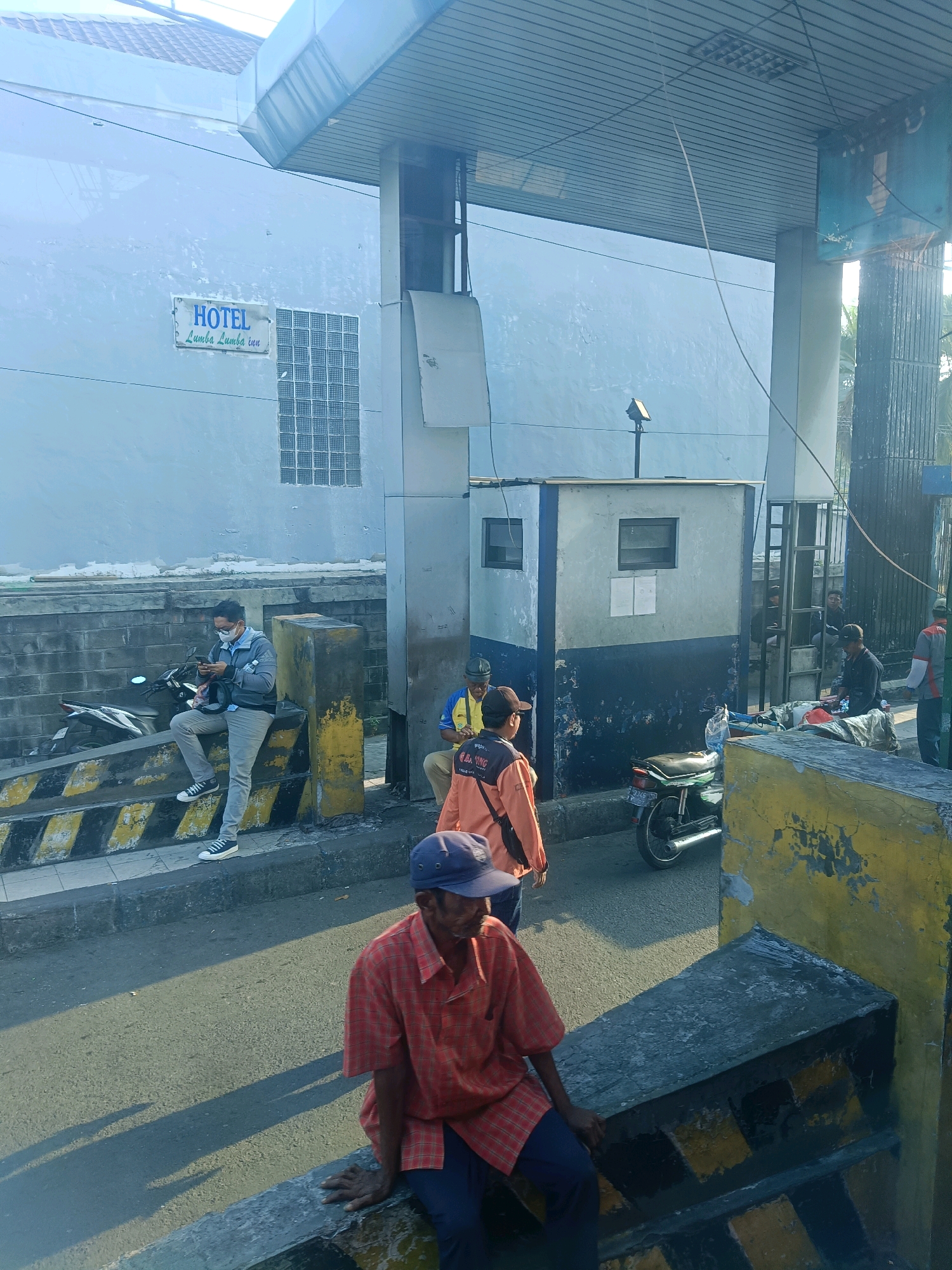 The image depicts an outdoor scene in Kedungrejo, Indonesia, likely near a toll booth or checkpoint. In the foreground, a man wearing an orange and red checkered shirt and a blue cap is sitting on a concrete barrier. Behind him, a motorcycle is parked. Further back, several other individuals are visible, some standing and some sitting near a small building with windows and a blue and white painted facade. A person in a grey jacket and face mask is sitting and looking at their phone. The building appears to be a service area or an office. There is a sign on the building that reads "HOTEL Lumba Lumba inn". The environment is urban, with a road and paved areas. The lighting suggests it is daytime, with shadows indicating the sun is present.