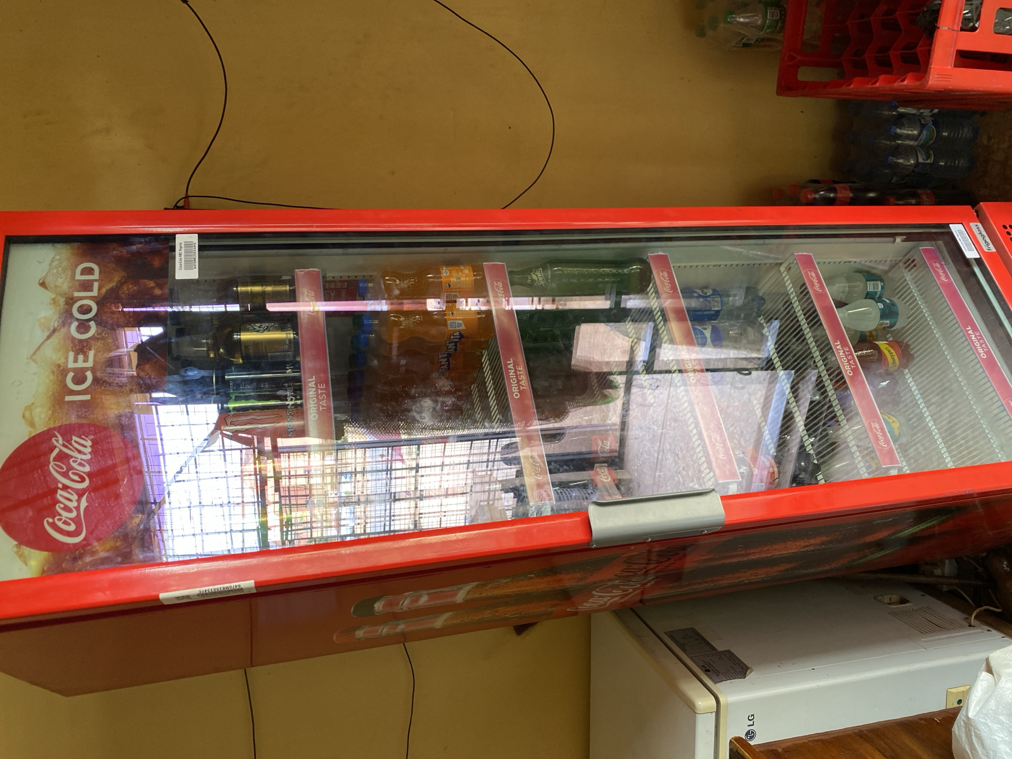 The image displays an indoor scene featuring a prominent red Coca-Cola branded display freezer. The freezer's glass door reveals an assortment of bottled beverages, including Coca-Cola, Fanta, Sprite, and clear water bottles, neatly arranged on shelves. Many bottle labels and shelf strips read "ORIGINAL TASTE," while the freezer's exterior bears the "Coca-Cola" logo and the text "ICE COLD."
To the right of the freezer, several red plastic crates are stacked, some filled with plastic water bottles. Below the freezer, part of a white appliance, possibly a refrigerator or air conditioning unit, is visible, featuring an "LG" logo. A wooden surface is also partially seen. A thin black wire is noticeable against the light yellow or beige wall in the background.
Visible text includes "Coca-Cola," "Fanta," "Sprite," "ICE COLD," and "ORIGINAL TASTE." A label on the freezer indicates "frigoglass," and another barcode label provides the text "0470ME25033410" and "Coca-Cola HBC Nigeria," confirming the regional distributor. This setup, with a commercial display freezer and beverage crates, strongly suggests a retail or storage area, likely a shop or kiosk in Bukuru, Nigeria. The image captures a static scene with no discernible human activity or specific time of day.