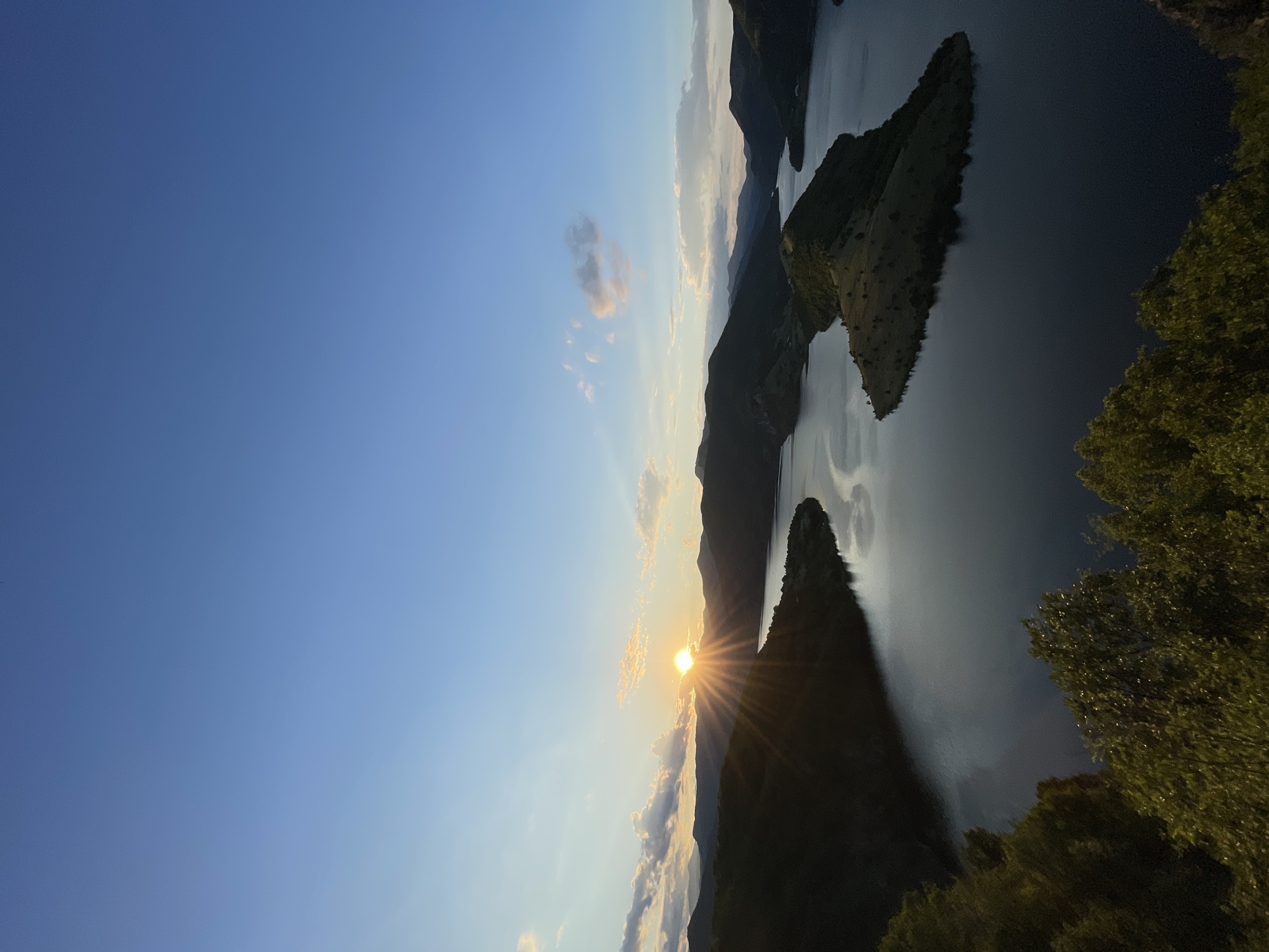 The image captures a serene sunset over a picturesque lake and mountainous terrain. The sun, partially obscured by the horizon, casts a warm, golden glow across the sky, creating a dramatic display of light rays that pierce through the clouds. The tranquil waters of the lake reflect the soft hues of the sunset, adding to the peaceful ambiance. Lush green foliage frames the foreground, adding depth and texture to the scene. The landscape appears to be the area around the city of Borovitsa, Bulgaria, known for its natural beauty and scenic vistas. No people or specific activities are depicted in the image, leaving the focus entirely on the breathtaking natural environment.