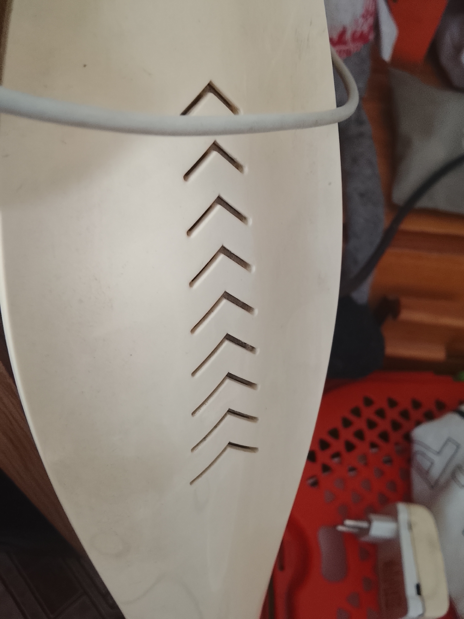 The image shows a close-up of a beige, oval-shaped object, possibly part of an electronic device or appliance. It has a series of V-shaped cutouts running vertically along its surface. A light grey cable is visible arcing over the top of the object. The background is out of focus but suggests an indoor setting with wooden furniture and some red and black patterned objects, as well as a white appliance with a plug. The lighting appears to be artificial. There is no visible text in the image. The scene is static, with no activity taking place. The image focuses on the texture and design of the beige object.