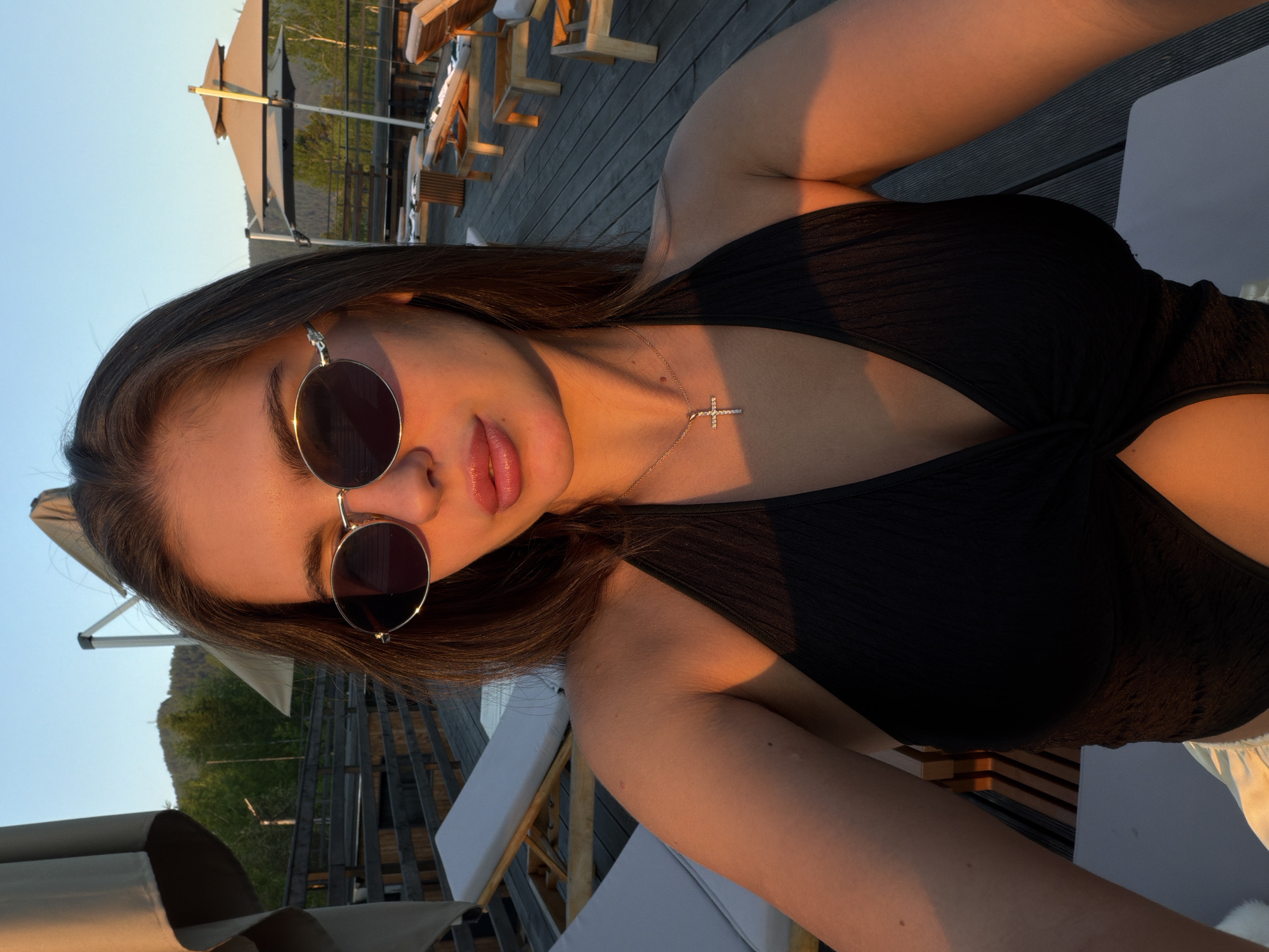 A young adult female is depicted in a close-up, self-portrait photograph taken outdoors during golden hour, presumably on a resort deck in Манский, Russia. She has dark brown hair and wears dark-lensed, oval-shaped sunglasses with silver frames. Her lips are full and appear to be enhanced with gloss or lipstick. She is wearing a black, textured halter-neck top with a front knot detail, exposing a portion of her midriff. A slender gold chain necklace with a small, jewel-encrusted cross pendant rests on her décolletage. Strong, warm, directional sunlight bathes her right side, imparting an orange tint to her skin and the top.
The background features a wooden deck with discernible planks, extending into the distance. Several empty lounge chairs with white cushions and light-colored wooden frames are arranged on the deck. Large, beige patio umbrellas, some open, are visible in various positions. Beyond the deck, a natural landscape of green trees and a distant hill or mountain is present under a clear, light blue sky.