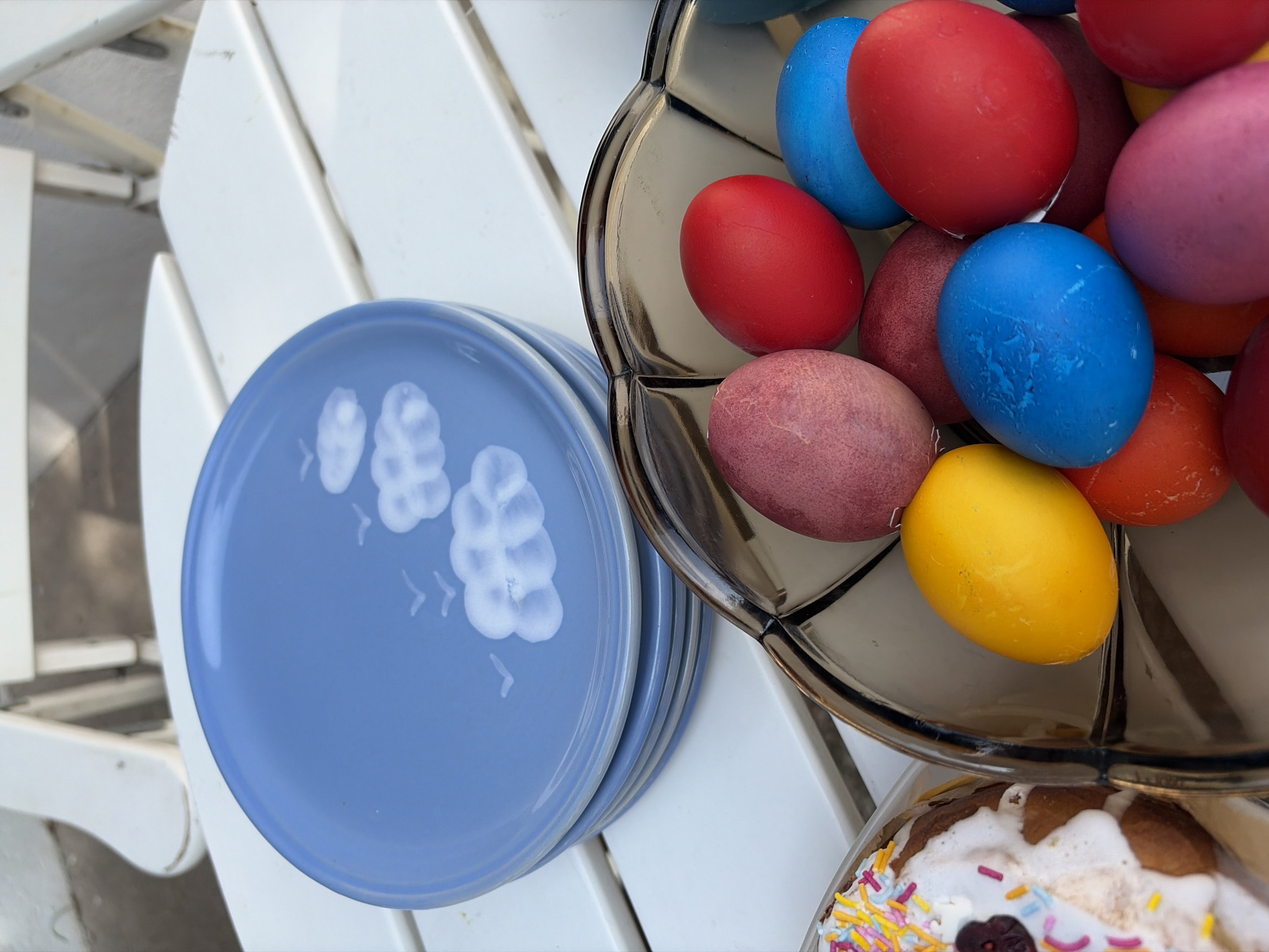 The image depicts a close-up of a collection of Easter items arranged on what appears to be a white outdoor table. To the left, there is a stack of light blue plates with a decorative white floral or leaf pattern. To the right, a clear, multi-tiered serving dish is filled with brightly colored Easter eggs. The eggs are painted in a variety of hues, including red, blue, yellow, orange, and pink. At the bottom right corner of the image, part of a cake or sweet bread, decorated with white frosting and colorful sprinkles, is visible. The scene is set outdoors in Topola Municipality, Serbia.