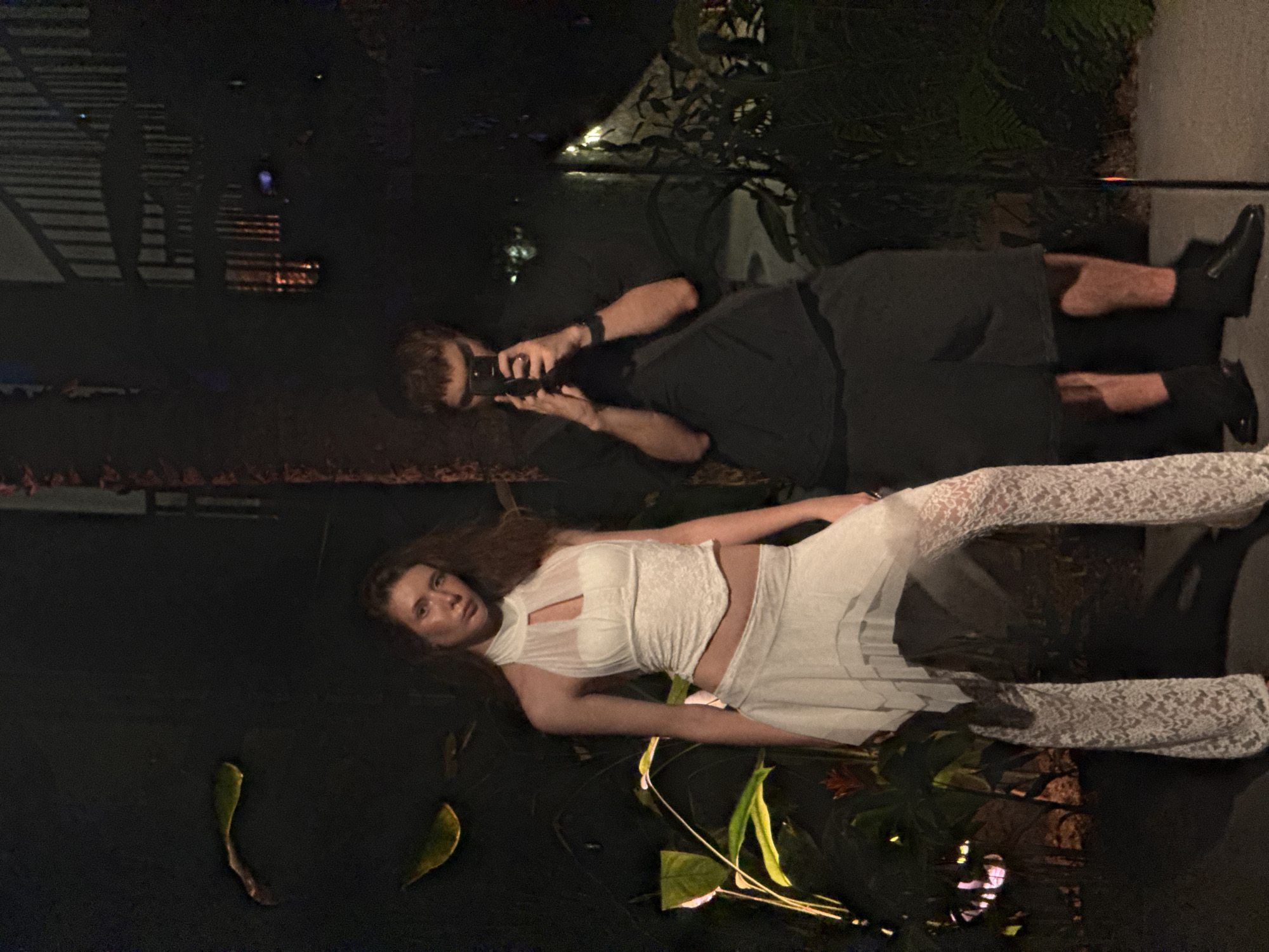 The image depicts a nighttime photoshoot featuring a man and a woman in what appears to be an urban or sophisticated architectural setting, possibly in Pudong, China. The scene is dimly lit by artificial light sources, creating strong reflections on a dark, reflective surface that resembles water.
The woman, with medium-length reddish-brown hair, is the model. She is lying on her back on the dark, reflective surface, looking directly towards the viewer with a neutral, focused expression. She is dressed in a striking white two-piece outfit: a high-neck, sleeveless top with sheer and textured fabric details, and matching white lace-patterned trousers or leggings. Around her, a few green leaves float on the dark surface.
Standing slightly behind and to the side of the woman is the man, who is the photographer. He has short brown hair and is wearing a dark t-shirt and dark pants with dark shoes. He is holding a black camera up to his face, indicating he is actively taking pictures of the woman.
In the background, reflections of distant artificial lights and architectural structures are visible on the dark surface, suggesting an urban environment. The time of day is clearly night, as indicated by the darkness and reliance on artificial illumination. There is no visible text in the image.