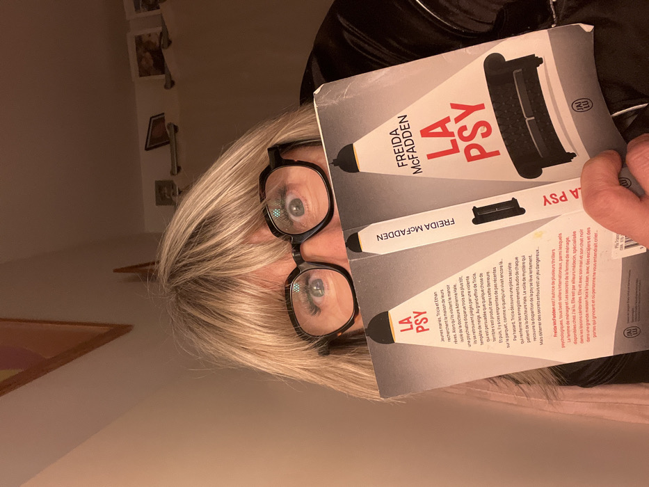 A woman with blonde hair and glasses holds up a book titled "La Psy" by Freida McFadden. The book cover features a black sofa and the author's name in bold. The back of the book describes the plot: A young couple, Tricia and Ethan, are trapped in a snowstorm at a mansion once owned by a missing psychiatrist, Adrienne Hale. Tricia discovers a secret room with audio recordings of Hale's patients, uncovering dangerous secrets. McFadden is an internationally bestselling author of psychological thrillers.