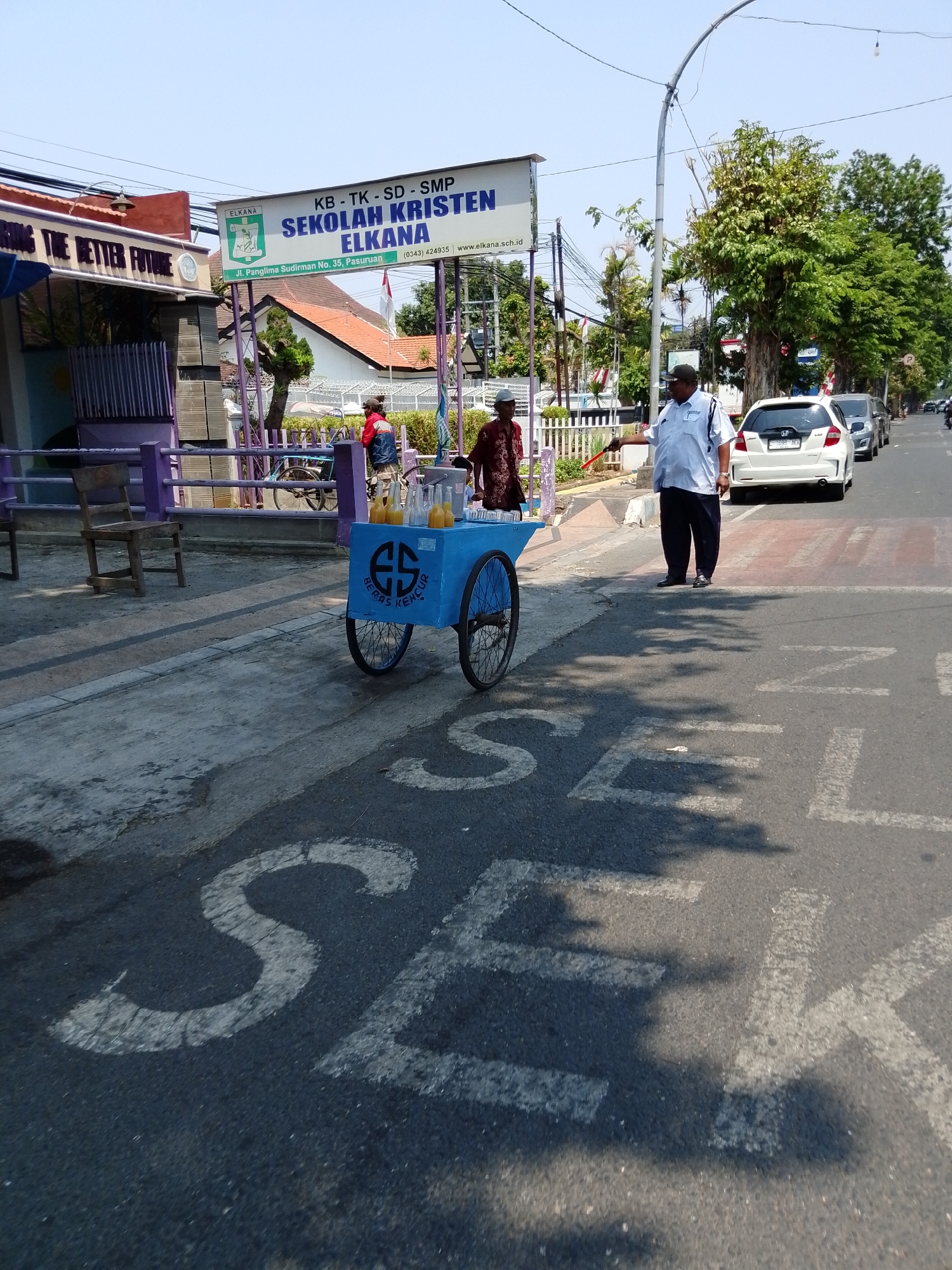 In Pasuruan, Indonesia, in front of the "Sekolah Kristen Elkana" school, a man in a white uniform, possibly a security guard or school staff, is directing traffic with a red stick. He is standing on the right side of the road, near a white car and a line of other vehicles. To his left, a man in a traditional garment is standing near a blue cart selling drinks, possibly "beras kencur," a traditional Indonesian beverage. The cart has the letters "ES" and "BERAS KENCUR" written on it. The scene is under a clear sky, with trees lining the street. The road has painted markings, including white letters that appear to spell "SEMUANYA" (meaning "everything" in Indonesian), suggesting a pedestrian crossing or a designated area. The school building has a prominent sign with the school's name and address.