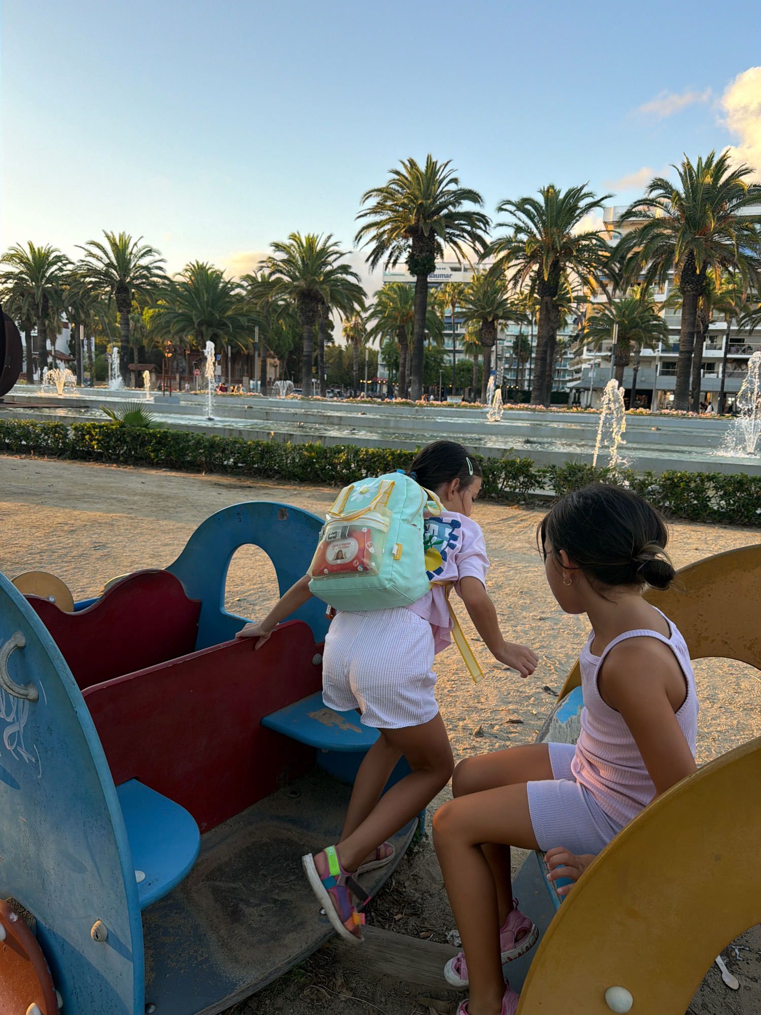 In Salou, Spain, two young girls are playing on a blue and red playground structure. The girl on the left, facing away from the camera, wears a light blue backpack with a graphic print and white striped shorts. She is reaching out with her right hand. The girl on the right is seated and faces the other child, wearing a light purple tank top and matching shorts. The playground is situated on a sandy surface, with a background of palm trees and fountains in what appears to be a park or public area. The lighting suggests it is late afternoon or early evening.
