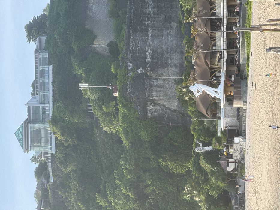 The image displays a multi-layered coastal scene in Ungasan, Indonesia. At the top, a modern, multi-story building with extensive glass facades and a prominent glass pyramidal roof sits atop a steep, densely vegetated green cliff. Below this, a cellular telecommunication tower with multiple antenna panels is positioned on a lower section of the cliff amidst the foliage. The cliff face exhibits visible, stratified rock formations.
In the middle ground, at the base of the cliff and adjacent to the beach, stands a multi-level building characterized by distinctive, undulating thatched or wavy roofs. This structure features open-air terraces where multiple individuals are seated at tables or lounges. A large, white statue of a female figure, depicted in flowing robes and holding an object resembling a stringed musical instrument, is positioned on a raised platform next to this building. To the left, a smaller, less distinct white humanoid statue with outstretched arms is visible, along with a traditional-style pavilion featuring a brown, peaked roof. Several palm trees are interspersed among these beachfront structures.
The foreground consists of a wide expanse of light brown sand, forming a beach. Multiple individuals are present on the sand: three people are grouped towards the left, appearing to walk or interact; one person walks centrally towards the right; and another individual, possibly a child, stands towards the right. Several weathered wooden posts or pilings are partially visible in the lower right corner of the beach. The sky overhead is clear and light blue.