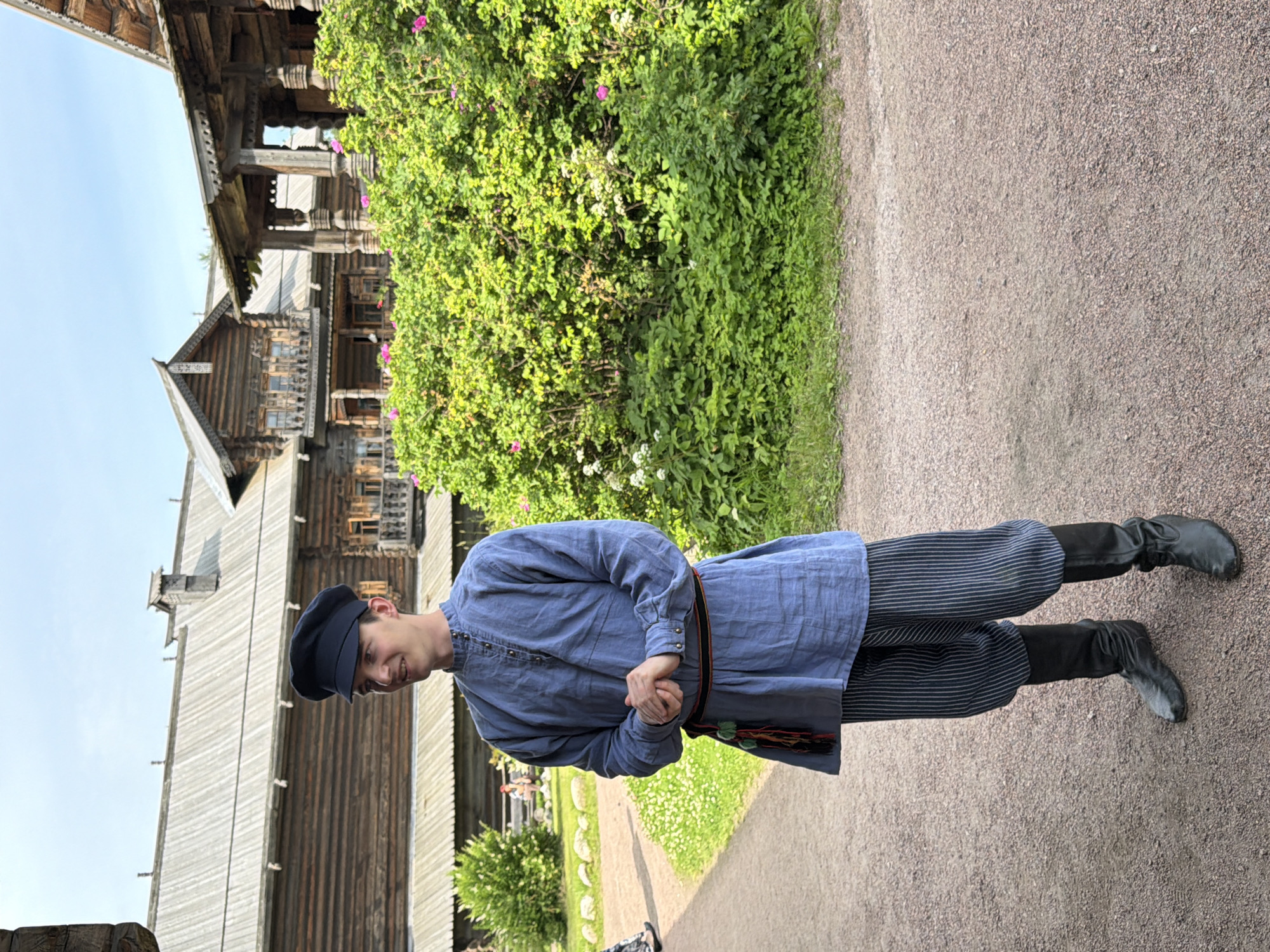 A young man stands outside in front of traditional wooden buildings. He is wearing a blue tunic with decorative buttons and striped pants. He also has on a dark cap and black boots. The buildings in the background are made of wood and have elaborate carvings. There is greenery and flowers surrounding the buildings. The ground is covered with gravel. The sky is clear and blue. The image appears to have been taken on a sunny day. The man's hands are clasped in front of him, and he is looking off to the side. He seems to be in a historical village or museum.