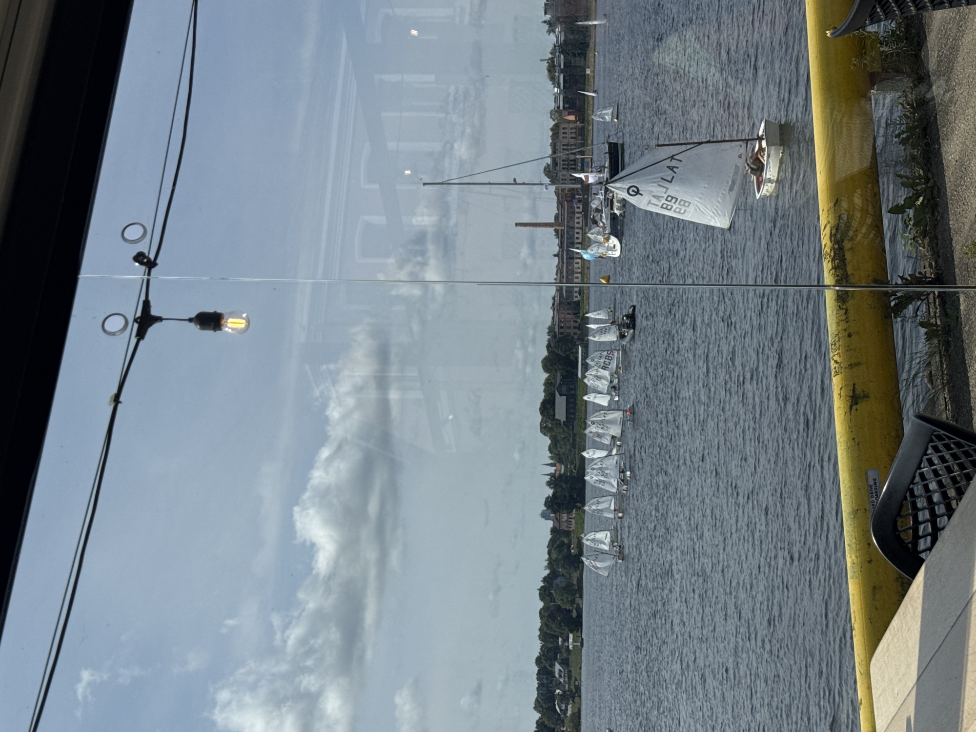 This image shows a wide shot of a body of water with numerous sailboats. The sailboats appear to be small dinghies, many of which are white with black markings and numbers on their sails. A larger sailboat with a tall mast is visible in the middle ground. The background features a shoreline with trees, buildings, and what appears to be an industrial chimney. The scene is framed by a window, with reflections of lights and the sky visible on the glass. The water is choppy, indicating a breezy day. The overall impression is of a sailing event or a regatta taking place.
Based on the visible markings on the sails of the sailboats, such as "LAT" and numbers like "89", and the context of a sailing event in a city with historical industrial architecture and a prominent chimney, this scene is likely located at the Daugava River in Riga, Latvia. Specifically, it could be near the Andrejsala or the area around the former Riga Shipyard, which often hosts sailing activities and has such distinctive industrial remnants. The presence of the yellow railing in the foreground suggests the view is from a waterfront establishment like a cafe or restaurant.