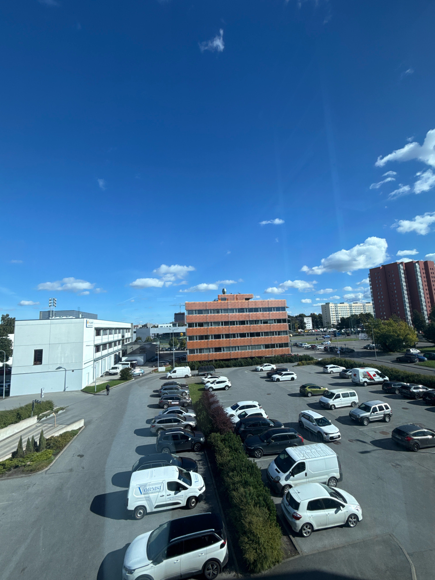 The image depicts a view of a parking lot and surrounding buildings in Tallinn, Estonia. The scene is dominated by a bright blue sky with scattered white clouds. Several vehicles are parked in the lot, including white vans, SUVs, and cars of various colors. Some buildings are visible, including an orange-colored office building and a taller, reddish-brown residential building. The parking lot appears to be located near an office or industrial area, with a modern white building on the left. A single person is walking in the parking lot. The overall impression is of a typical urban environment on a sunny day.