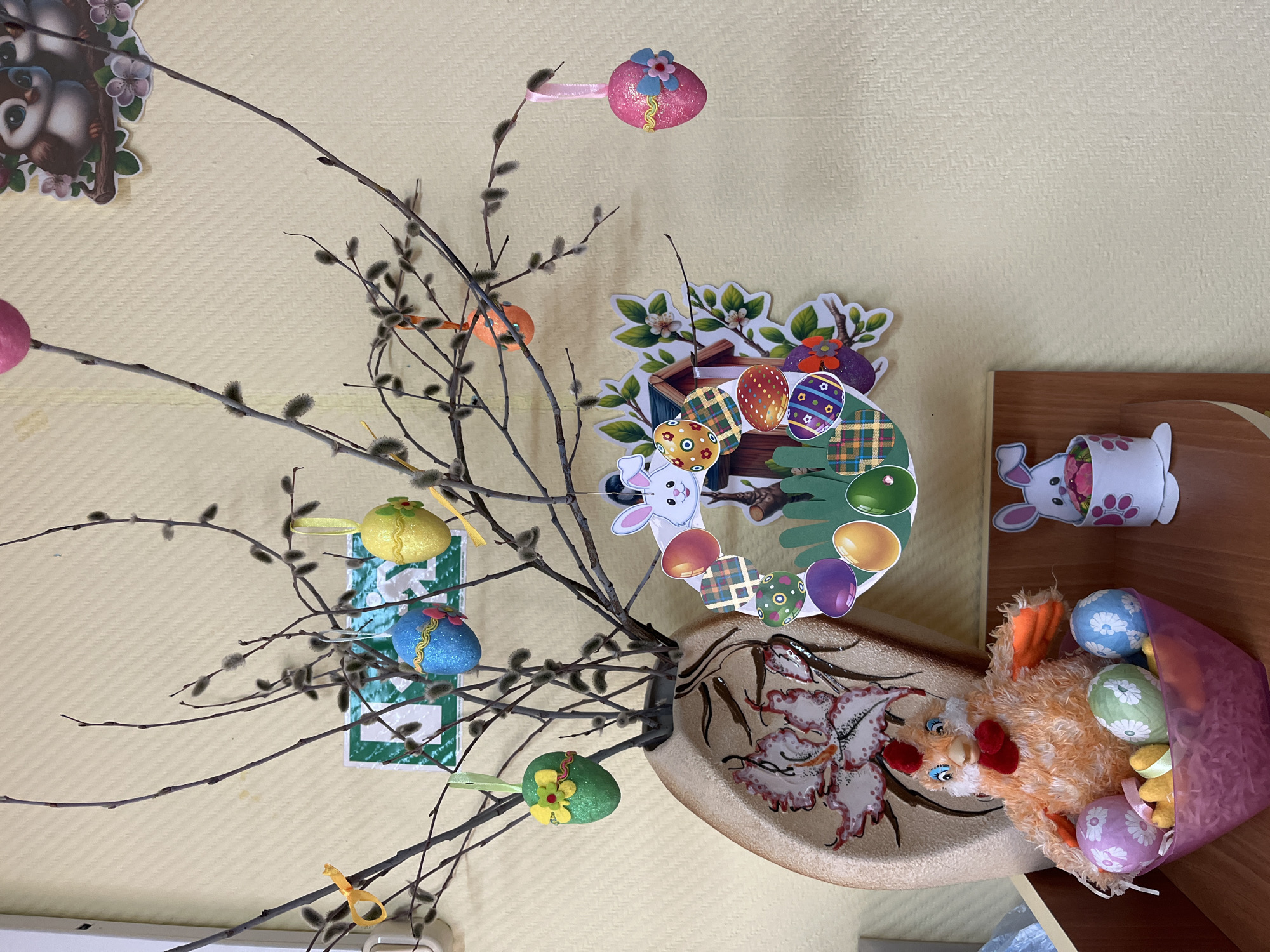 The image depicts an Easter-themed arrangement, likely inside a building, given the textured yellow wall in the background. A vase or bowl with a floral design holds branches of willow, decorated with Easter egg ornaments in varying colors and designs (glittery, floral, etc.). Easter-themed cutouts and stickers (rabbits, egg-filled baskets, wreaths) are placed around the arrangement and on the wall. A stuffed chicken toy sits near the vase, with a small, purple bag overflowing with colored eggs. A wooden shelf is partially visible. Based on the metadata, the location is Chaikovsky, Russia. No people are visible in the scene. The interaction is between the objects and the placement of the objects. The scene is festive.