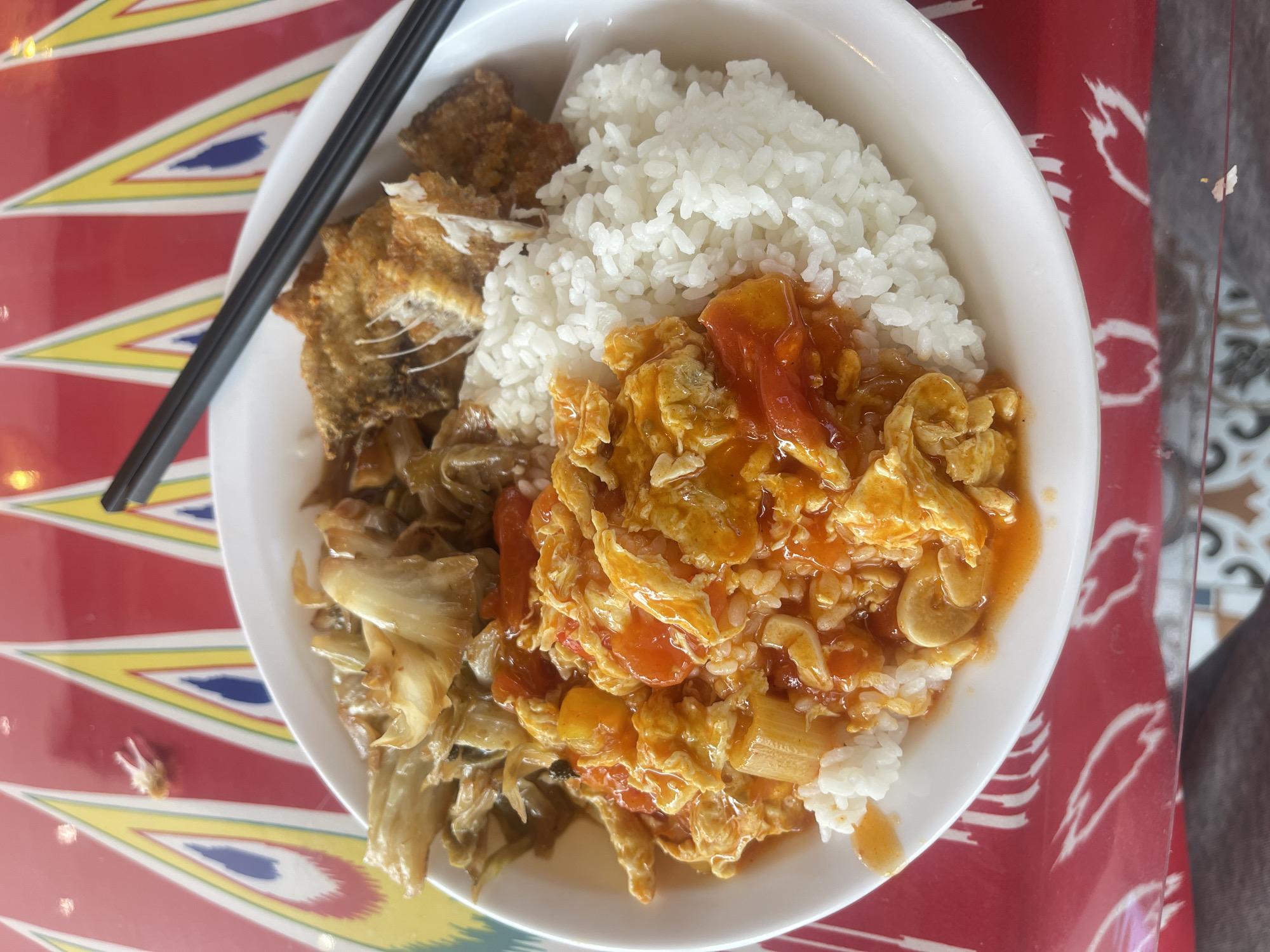 A white bowl of food sits on a table in what appears to be Chongqing High-tech Zone, China, based on the style of the tablecloth. The bowl contains rice, fried fish, a vegetable dish (possibly cabbage), and a tomato and egg dish. Black chopsticks rest on the edge of the bowl. The tablecloth is red and white with a yellow and blue geometric pattern.