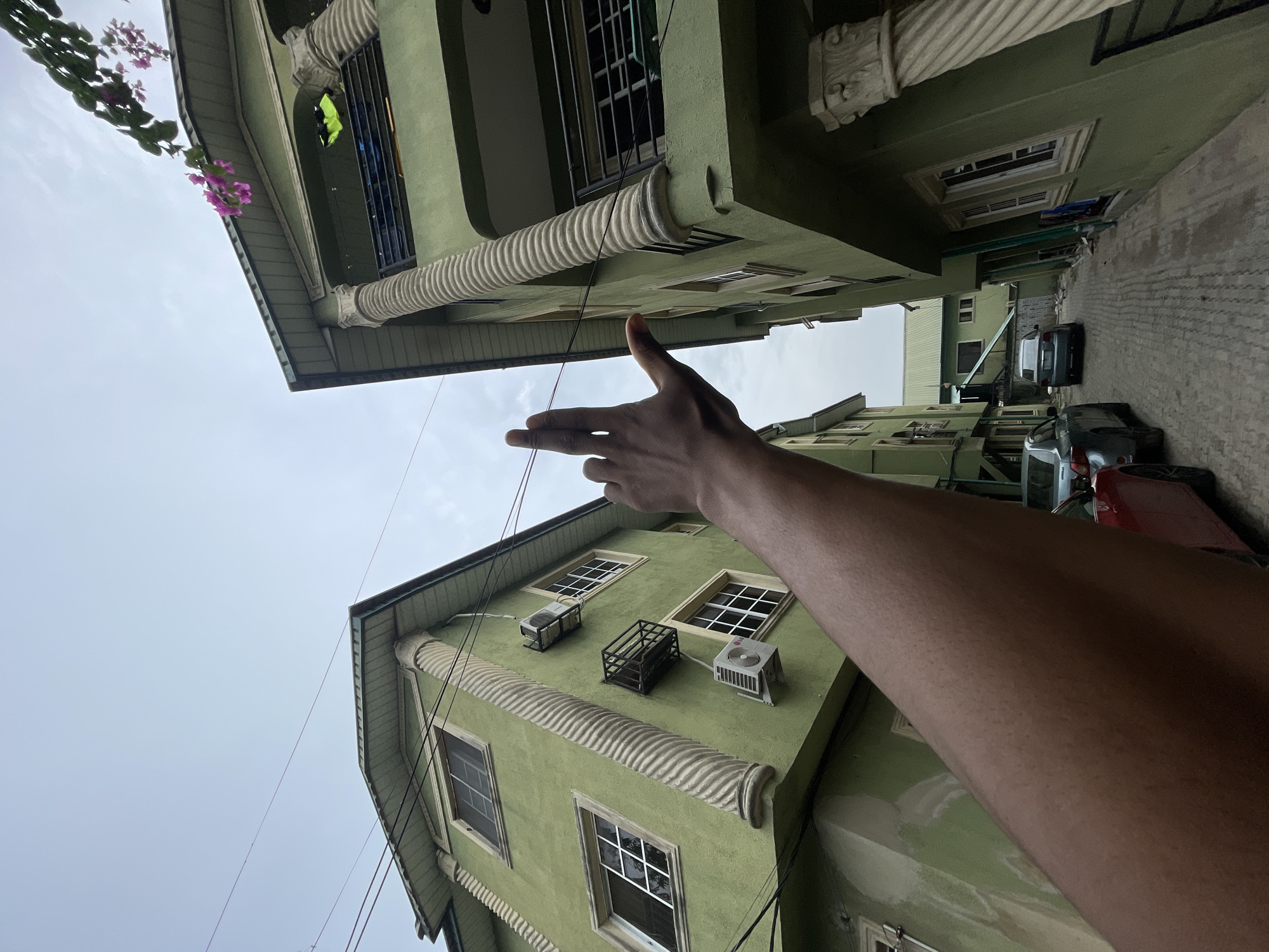 The image, taken in Ado, Nigeria, presents a low-angle view of two buildings painted in a similar shade of light green. The buildings are situated close to each other, forming a narrow alleyway visible towards the top right of the frame. Several cars are parked along the side of the alleyway, including a silver or gray vehicle and a red vehicle. The buildings feature decorative moldings, multiple windows, and what appears to be air conditioning units mounted on the exterior of the lower building. An arm extends upwards into the frame, with fingers pointed towards the building above. The sky is overcast and gray, without any notable features. A power cable runs diagonally across the view. Some pink flowers are seen at the top of the building in the top left.