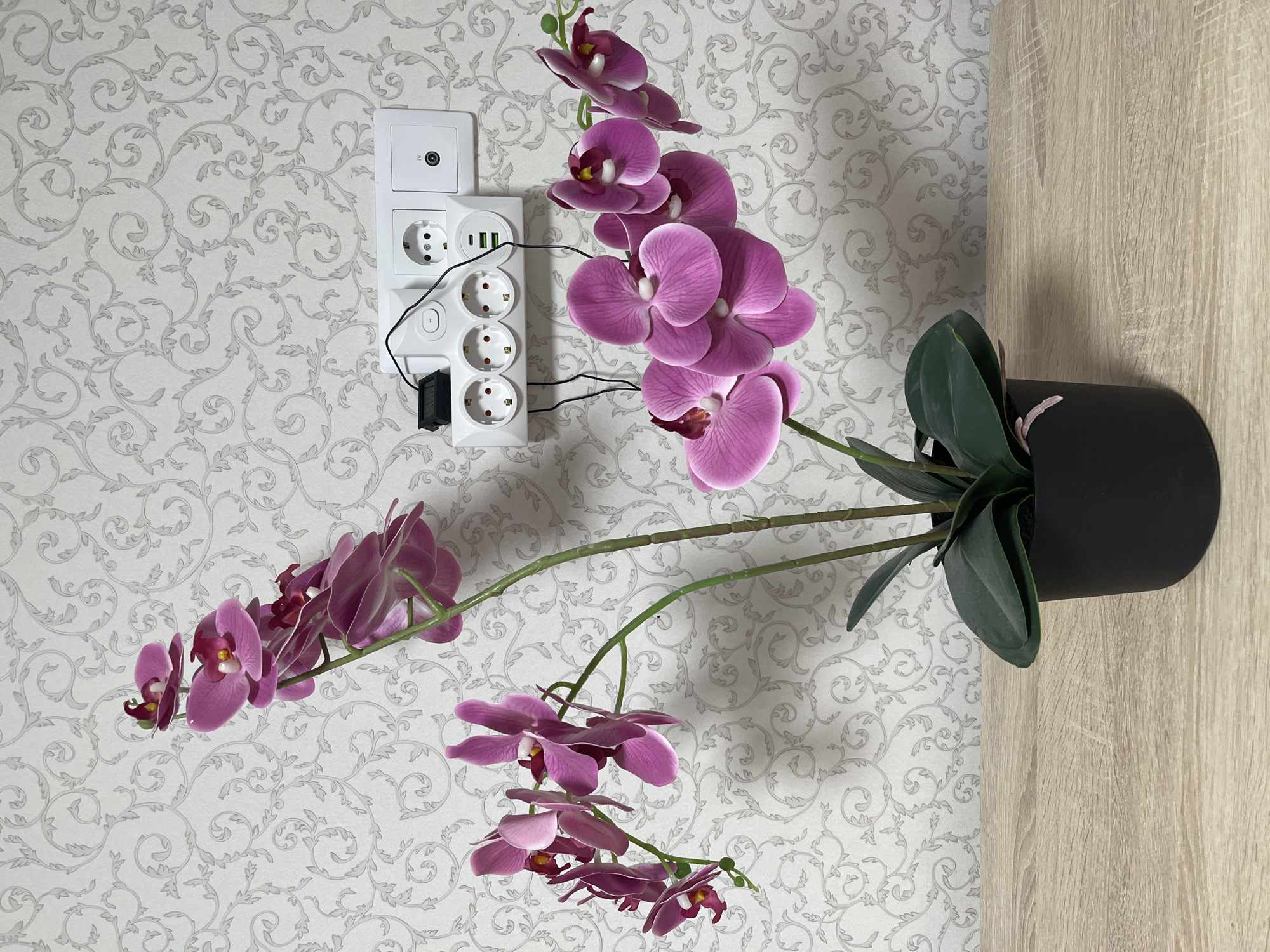 This detailed photograph, captured in Старые Кены, Russia, presents an indoor scene featuring an artificial potted orchid. The orchid, positioned in the lower-center, has multiple branches adorned with numerous vibrant purple flowers, each with a white and yellow center, and several dark green leaves. It is planted in a simple black, cylindrical pot. The pot rests on a light-colored wooden surface exhibiting a distinct grain pattern. The background consists of a light off-white or light gray wallpaper patterned with repeating, intricate silver or light gray floral and scroll motifs. Mounted on the wallpaper, to the upper-right of the orchid, is a white electrical power strip with three European-style outlets and two green USB charging ports. A small black digital display device, likely a USB voltage/current tester, is plugged into the top-left outlet of the power strip, with a thin black cable extending from it. To the immediate right of the power strip, a separate white wall plate contains one European-style outlet and one coaxial 'TV' port. The scene is evenly lit.
