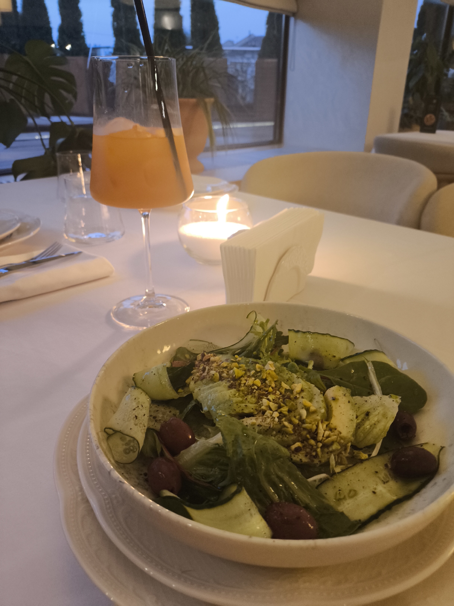 Here is a detailed description of the image:
The image shows a close-up view of a restaurant table setting, likely in the evening. The table is covered with a white tablecloth, and several items are arranged on it.
In the foreground, a white bowl filled with a salad dominates the view. The salad appears to contain a mix of lettuce, cucumbers sliced into roll shapes, olives, and a topping of chopped nuts, possibly pistachios. The bowl is placed on a white plate.
To the left of the salad, there is a set of silverware neatly placed on a napkin. Behind the salad bowl, a glass of orange juice with a black straw stands prominently. Next to the juice, a small candle in a glass holder emits a soft light, and a stack of white napkins is also visible.
Further back, the background shows a blurred view of the restaurant interior. One can discern the presence of beige or cream-colored chairs, a window with a partial view of what appears to be city lights at dusk, and some greenery (possibly potted plants) near the window. The presence of evening lighting and partially visible building structures indicate an urban setting but the city location is unknown.