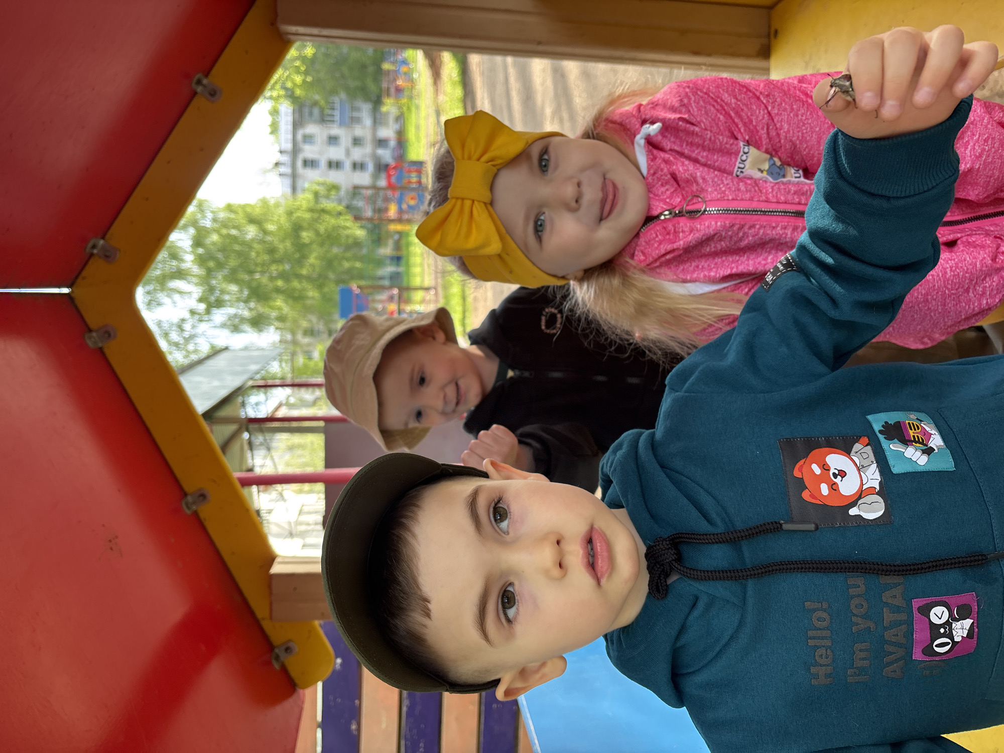 A medium shot depicts three young children playing on a colorful playground structure. The dominant child in the foreground, a boy wearing a dark green cap and a blue-green hooded sweatshirt with cartoon patches, looks directly at the camera. Behind him and slightly to the left, another child, wearing a tan bucket hat and a black jacket, smiles broadly, partially obscured. To the right of the boy in the cap, a girl wearing a pink hooded jacket and a large yellow bow in her hair is sticking her tongue out and holding an insect on her fingertip.
The playground's structure consists of red, yellow, and blue elements. The backdrop shows trees, buildings, and a playset suggesting the outdoor setting. Given the city of Chaikovsky, Russia as the location, the image likely represents children at play in a public park or playground within the city. The children are interacting with their environment and each other as they engage in play.