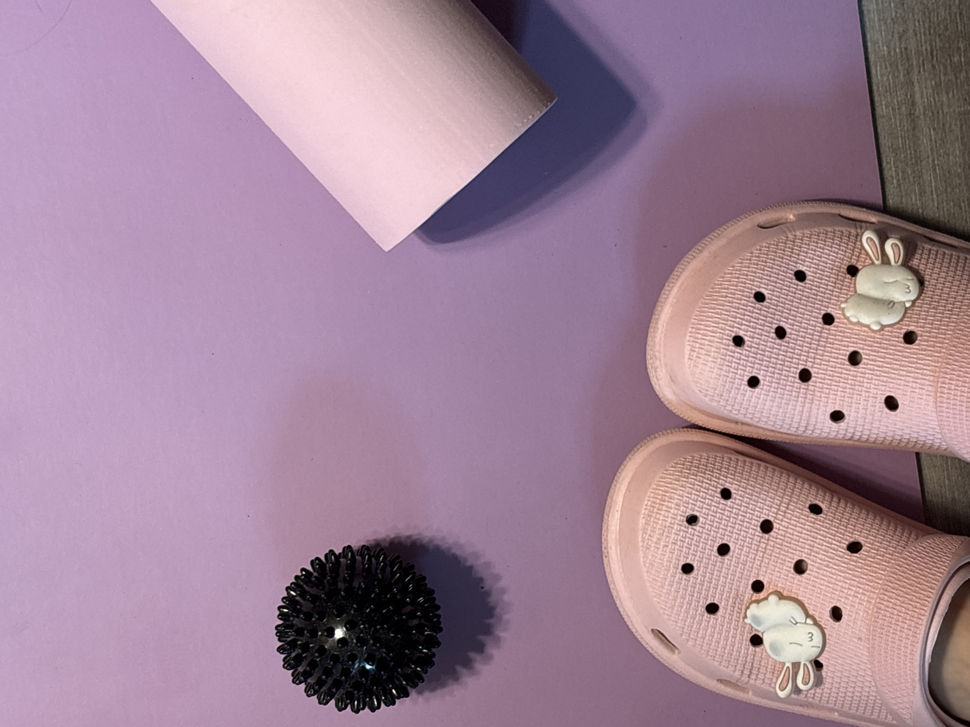 The image presents a flat lay composition on a light purple surface, possibly a yoga mat. In the upper left quadrant, a light pink foam roller lies diagonally. A spiky black massage ball is positioned in the lower left quadrant. In the lower right quadrant, a pair of light pink Crocs-style shoes are placed, each adorned with a white bunny charm. A grey wood-patterned surface borders the mat on the right side. The scene lacks any indicators related to Saint Petersburg, Russia.