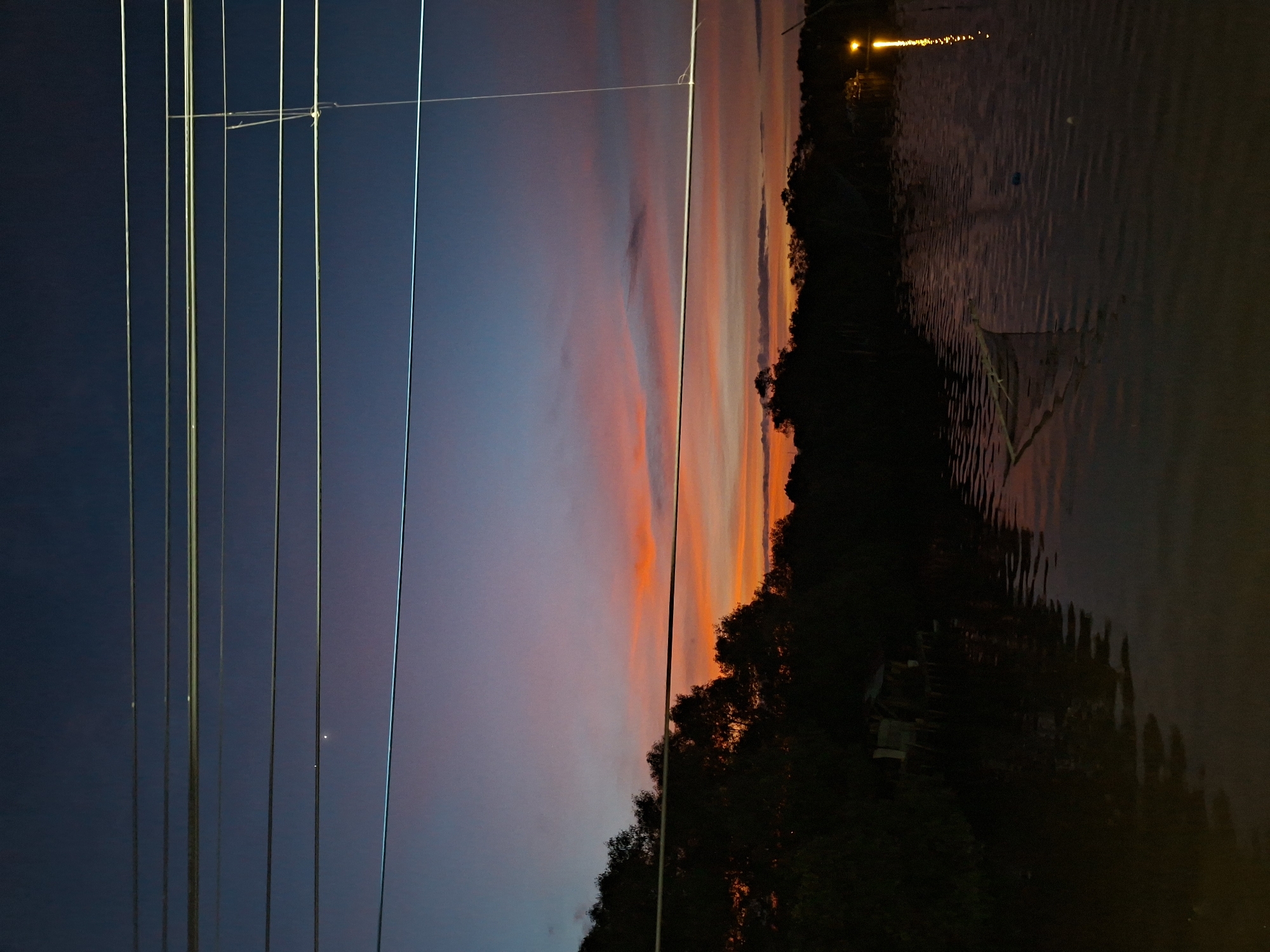 This is a dusk scene over a body of water, possibly a river or bay, in Lhokseumawe, Indonesia. The sky displays a gradient of deep blue at the top, transitioning through shades of purple and pink to a fiery orange band just above the horizon. Silhouetted against the colorful sky is a dark line of trees and vegetation that forms the coastline.
In the foreground, the water is dark and reflective, with ripples distorting the reflections of the sky and the surrounding environment. A small, possibly abandoned, structure built on stilts is visible on the left side of the image, partially submerged in the water and surrounded by dense foliage. Further to the right, a white fishing net or trap floats on the surface of the water, and a single light source illuminates a reflection on the water, suggesting a dwelling or activity on the distant shore.
Several utility wires stretch horizontally across the upper portion of the image, bisecting the sky and obscuring a small, bright point of light, possibly a star or planet, in the upper left quadrant. The overall atmosphere is tranquil and serene, capturing the fading light of day. There are no people visible in the image.