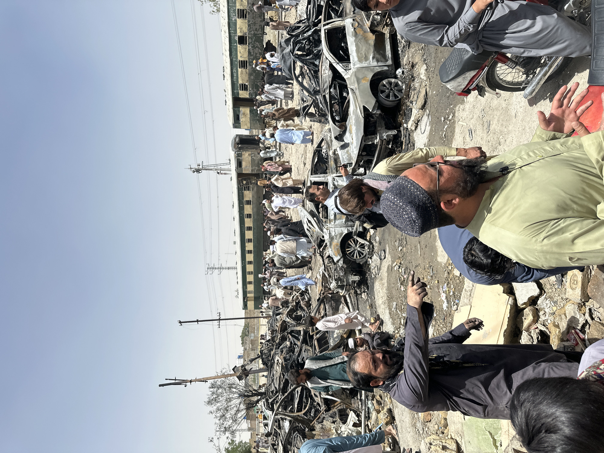 The image depicts the aftermath of an apparent disaster scene in Quetta City Tehsil, Pakistan. The scene is filled with debris, including the charred remains of multiple cars. A passenger train with green cars is parked nearby. A crowd of people, mostly men dressed in traditional Pakistani clothing, are present. Some appear to be examining the damage, while others are observing the scene. Power lines and utility poles are visible in the background. The environment appears to be a roadside or open area adjacent to a train track.