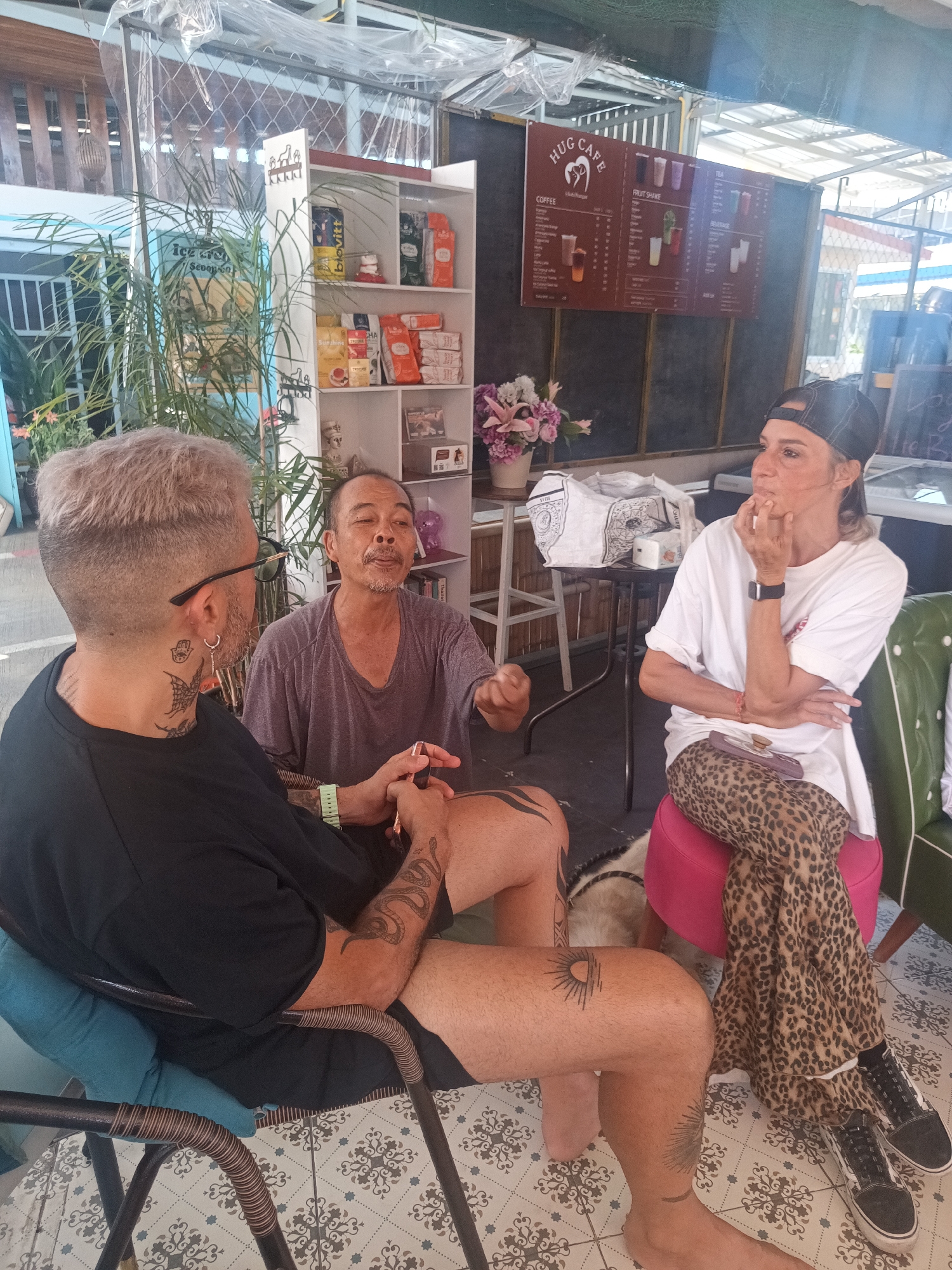 The image shows three people in what appears to be an outdoor cafe setting, possibly in ตำบลเกาะพะงัน, Thailand, given the prompt. A person with dyed blonde hair and numerous tattoos is seated in a blue chair, facing the camera at an angle. Next to them, a person with short, graying hair is seated and gesturing with their hand, possibly speaking. Across from them, a person wearing a white t-shirt, leopard-print pants, and a baseball cap sits on a pink ottoman, with their hand to their chin as if in thought.
In the background, a white bookshelf with various items and a menu board labeled "HUG CAFE" are visible. There is a small table with a flower arrangement and a white bag. The floor has a patterned tile design. The cafe is partially covered by a transparent awning.