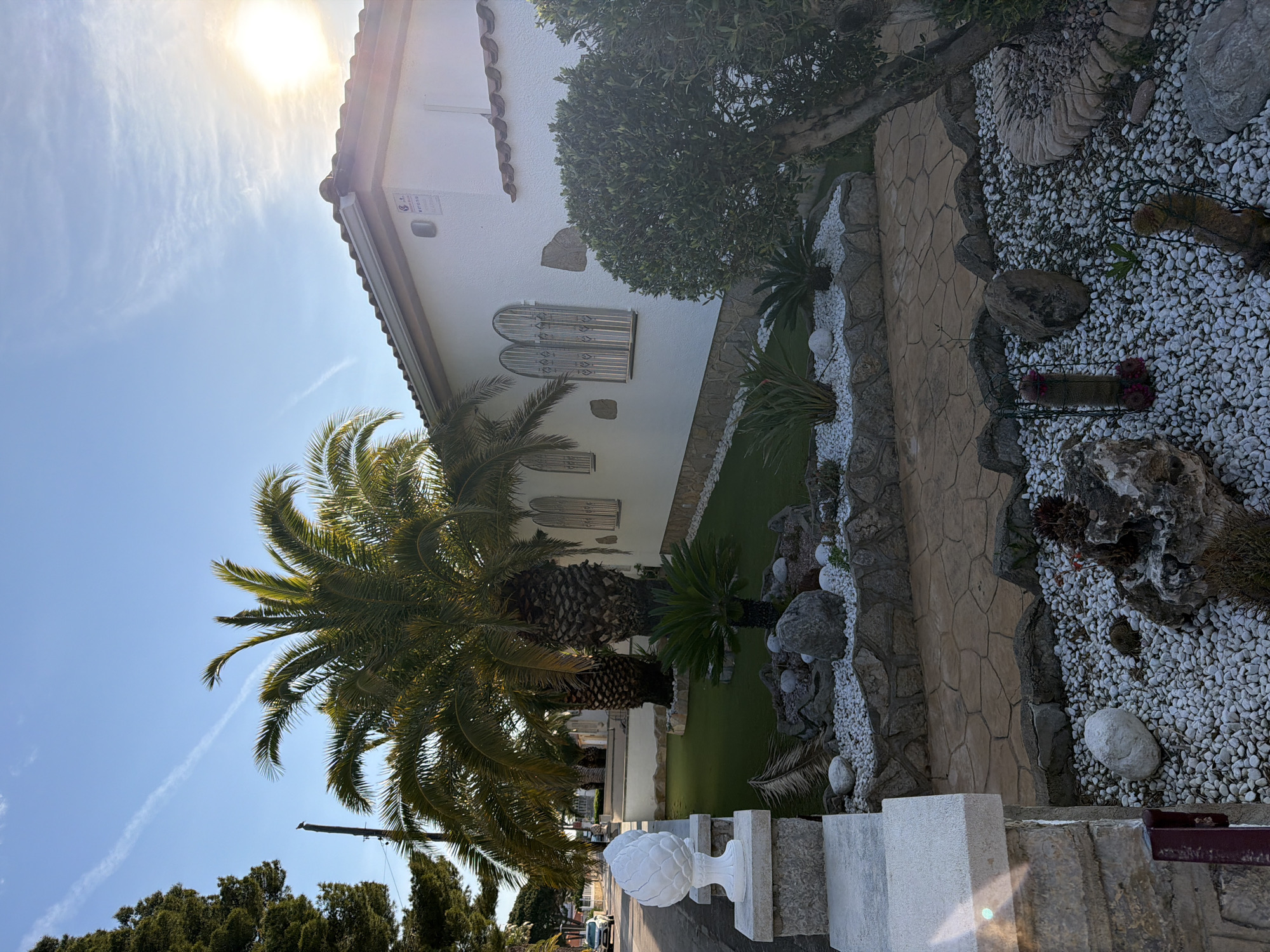 The image shows a house and garden in Mont-roig del Camp, Spain. The house is white with a brown tiled roof and has several arched windows with security bars. A large palm tree is in the front yard, along with other smaller trees and plants. There is a garden area with white stones and a stone pathway. A decorative stone post stands near the sidewalk. The sky is blue with a few clouds and the sun is shining brightly.