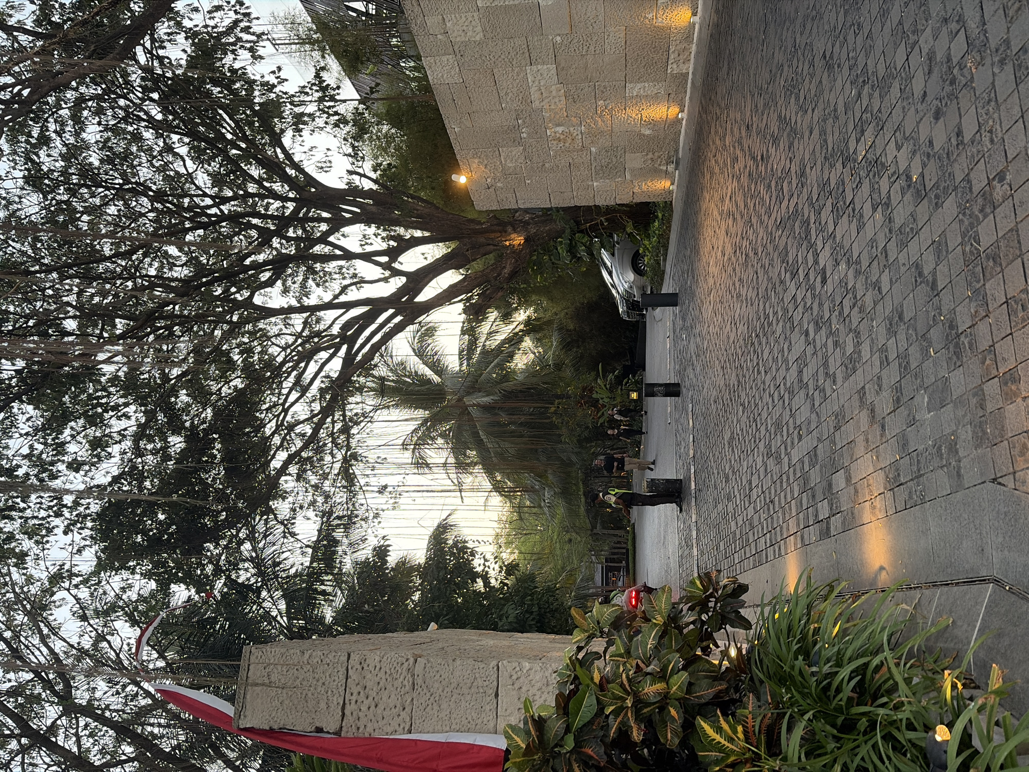 A paved driveway leads into a resort or hotel in Kuta, Indonesia, at dusk. The driveway is made of dark grey interlocking pavers. To the left, a light-colored stone pillar is adorned with a red and white Indonesian flag. Lush green plants with variegated leaves grow along the edge of the driveway.
In the background, several people are walking on the driveway. One person, wearing a high-visibility vest, stands near a bollard. Further down the driveway, two cars are parked. The area is illuminated by strategically placed orange lights embedded in the wall on the right and at the base of the driveway, casting warm glows.
Towering trees with dense foliage and hanging vines dominate the upper portion of the image. Palm trees are also visible, suggesting a tropical environment. The sky is overcast, indicative of sunset or twilight. The overall scene depicts an entrance to a well-maintained establishment in a tropical urban setting.