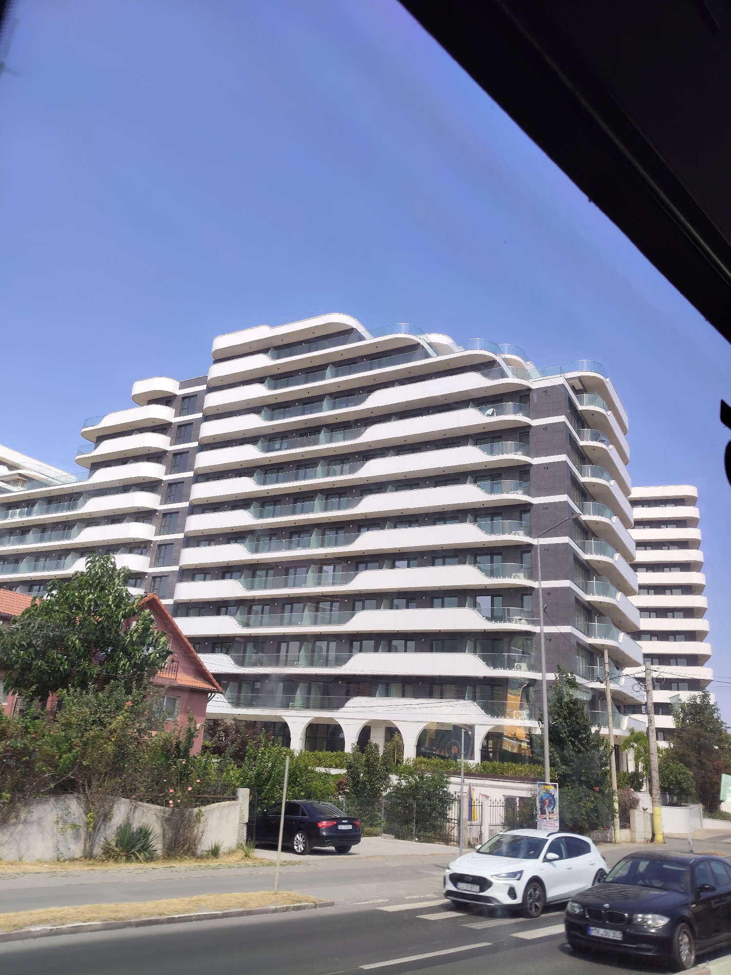 In a city scene under a clear blue sky, a modern, multi-story apartment building with a distinctive wave-like facade dominates the background. The building features white curved balconies with glass railings and dark-paneled sections. To the left, a smaller, older, reddish-brown house with a gabled roof is partially visible, surrounded by lush green trees and bushes. In the foreground, a street runs horizontally across the image. A black sedan is parked on the right side of the street. Further down the street, two cars are driving on a crosswalk: a white hatchback in the left lane and a black sedan in the right lane. The environment suggests a sunny day.