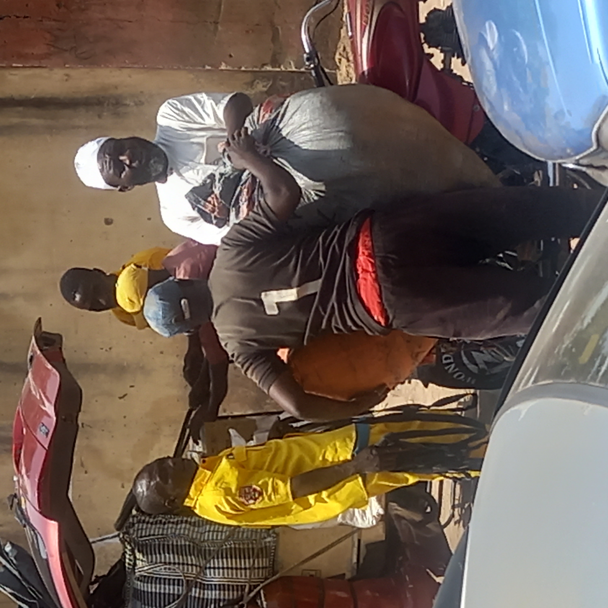 The image shows four men and two motorbikes laden with bags, presumably in the city of Dadi Riba, Nigeria. One man wears a white robe and cap, another wears a yellow t-shirt, one wears a black shirt with a white number "7" on it and a blue cap, and the fourth wears a yellow shirt. The bikes are red and blue.