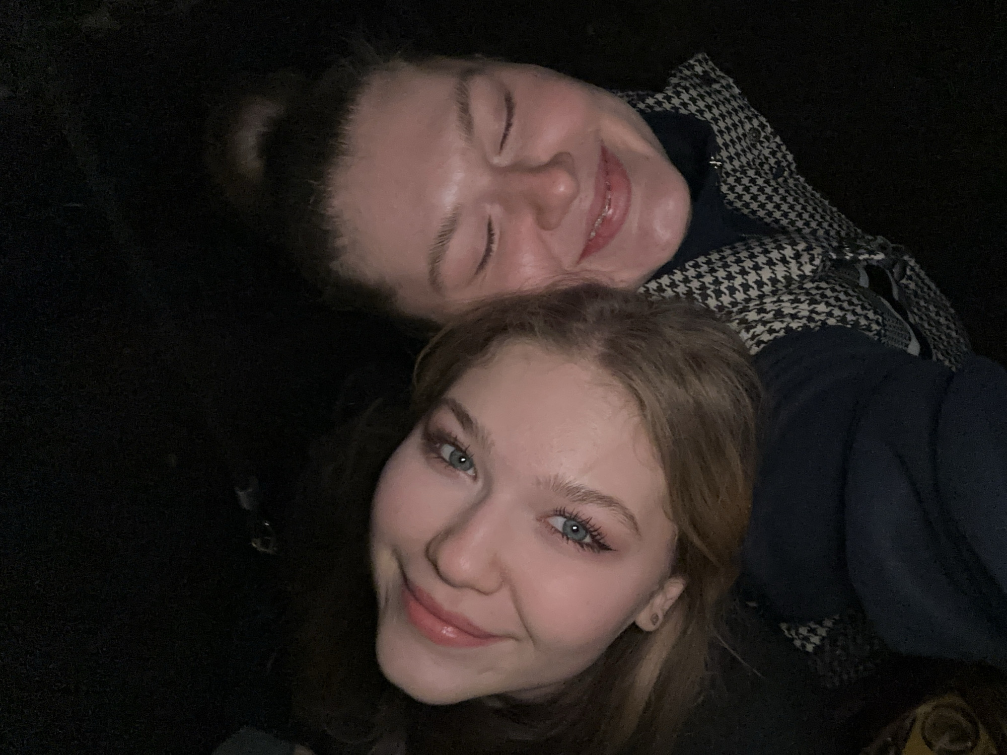 Two young women are shown in a close-up selfie. The woman in the foreground has blonde hair, fair skin, blue eyes, and wears pink lip gloss. She looks directly at the camera and smiles. The woman behind her has her eyes closed and is also smiling. She is wearing a houndstooth patterned coat. The background is dark and indistinct. It is not possible to confirm the location as Kazan, Russia based on the provided visual information.