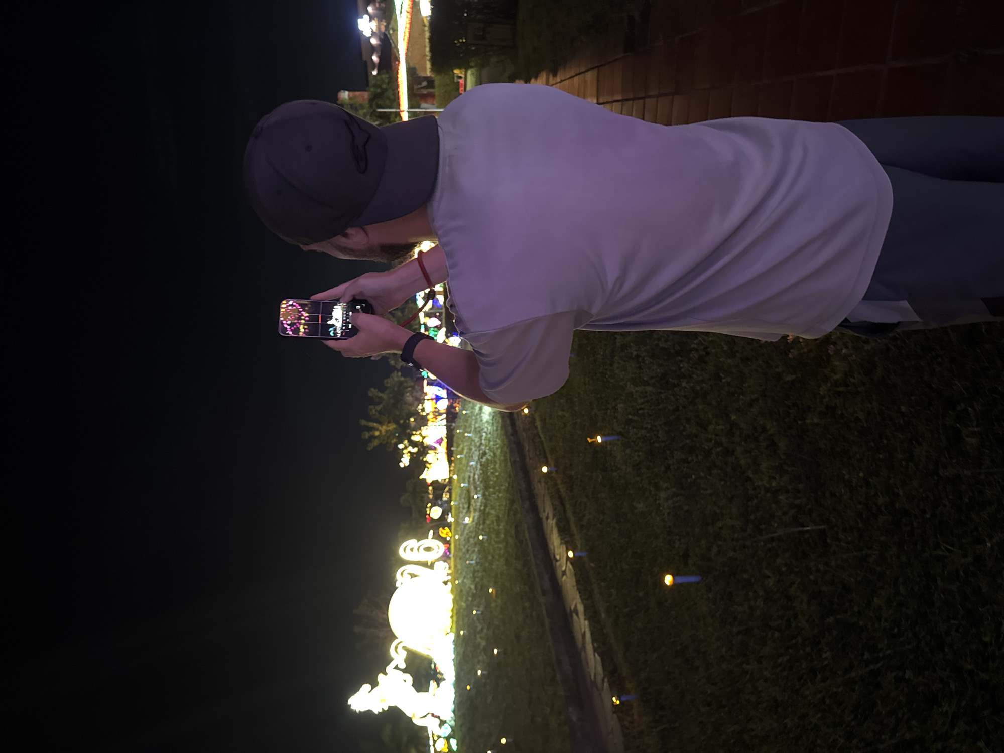 At night in Hội An Ward, Vietnam, a person wearing a backwards baseball cap and a short-sleeved t-shirt is taking a photograph with a smartphone of a light display, likely part of a seasonal celebration or festival, on a grassy area, with small light posts marking a pathway. The path appears to lead further toward the light installations.