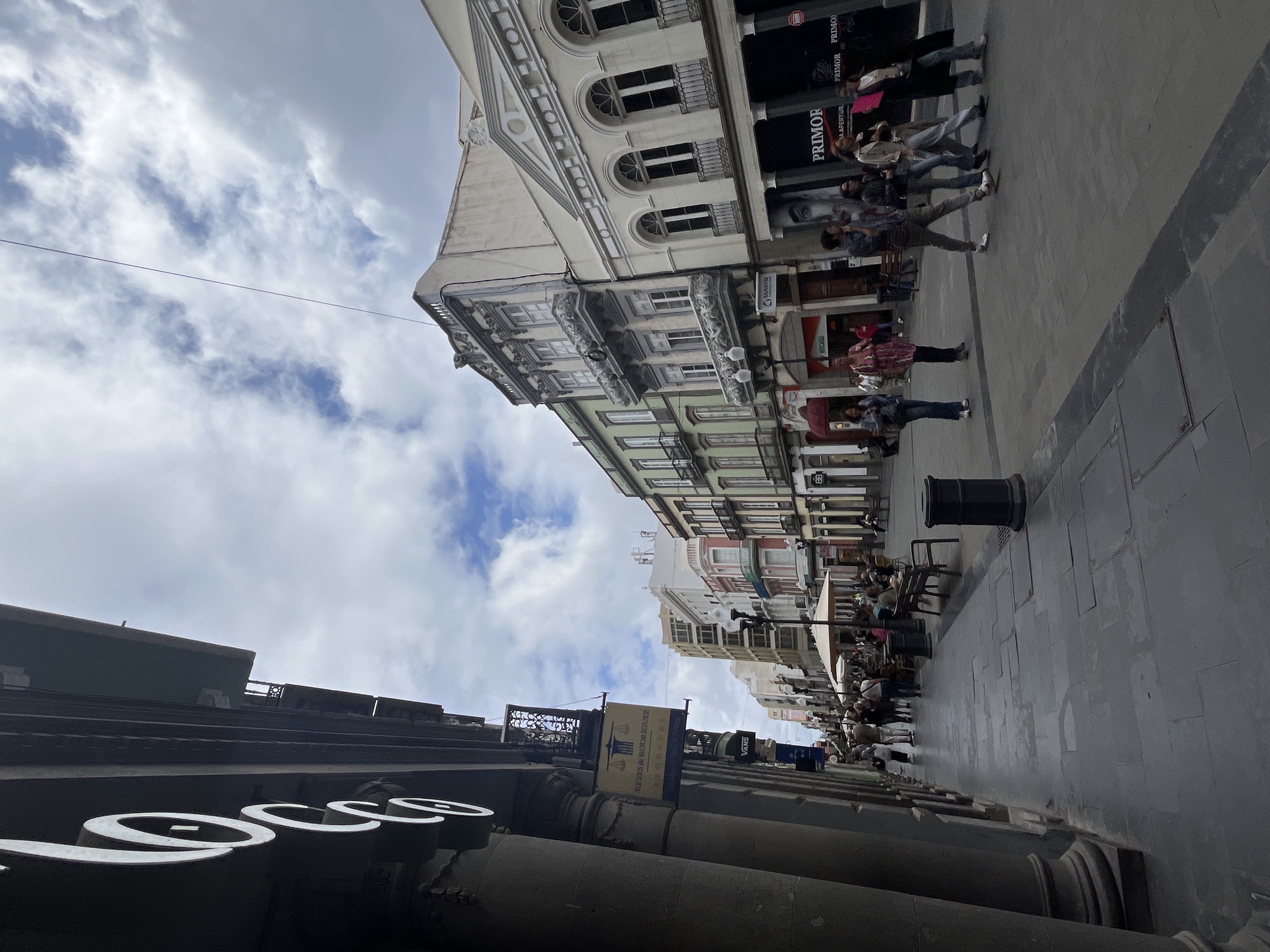 The image, correctly oriented, captures a vibrant daytime street scene in what appears to be a city in Las Palmas, Spain. The view is from a low angle, looking up a paved pedestrian street flanked by multi-story buildings under a partially cloudy sky.
On the right side of the street, a row of architecturally distinct buildings lines the thoroughfare. The closest building boasts a light-colored, classical facade with arched windows, decorative balconies, and ornate cornices. Further down, buildings exhibit a mix of styles, including one with a mint-green exterior. A prominent dark storefront displays the large white text "PRIMOR" and the partial phrase "LA APERTUR." Numerous people are visible along this sidewalk; some are walking, while others are seated at outdoor tables belonging to cafes or restaurants, some under umbrellas.
The left side of the street features a darker, more robust building with large, circular columns at its base. Prominent stylized white letters, possibly forming part of a word such as "ESSE" or "LOES," adorn its facade. A blue and yellow banner, suspended between buildings, clearly reads "EYES & RODRIGUEZ" and "ABOGADOS" (Lawyers), accompanied by a scales of justice logo. A small "VANS" logo is also discernible on a sign. The sidewalk here is wide and paved with gray blocks, furnished with a black waste bin and benches.
The sky above is bright, with scattered white clouds and visible patches of blue. The scene depicts typical daily urban life, with people engaged in walking, shopping, and dining, suggesting a lively and active atmosphere. The visible Spanish text on banners and storefronts serves as a clear location cue for a Spanish-speaking region.